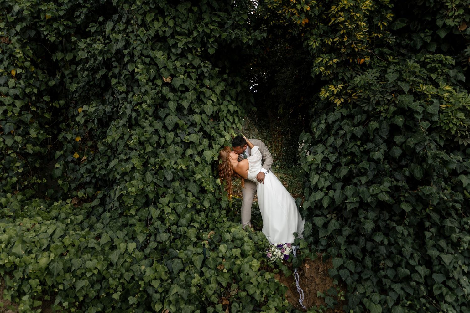 Couple sharing a romantic kiss surrounded by lush green ivy wall in an outdoor wedding photo.