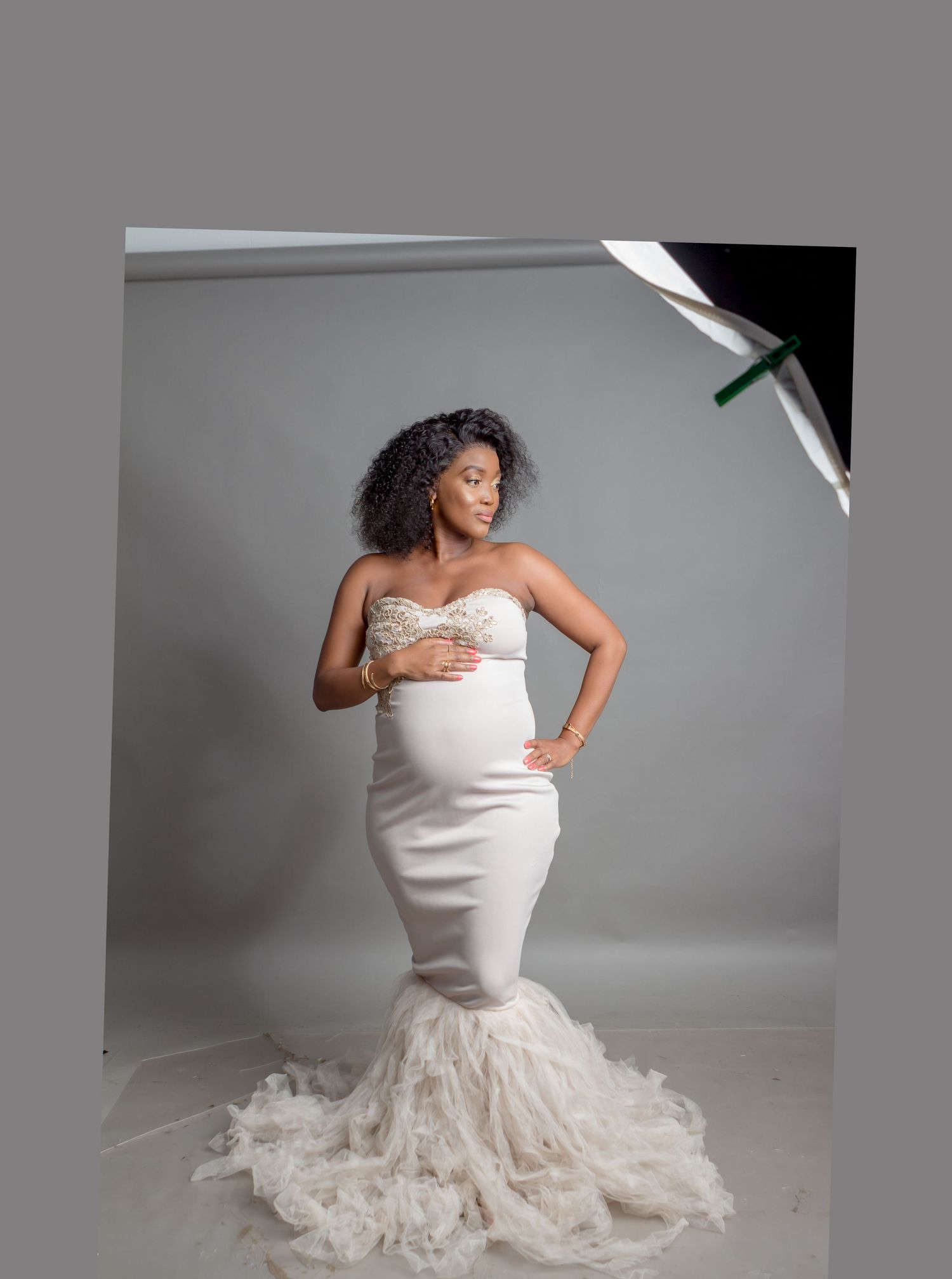 A pregnant woman poses in an elegant white mermaid-style gown with feathered train during a maternity photoshoot.