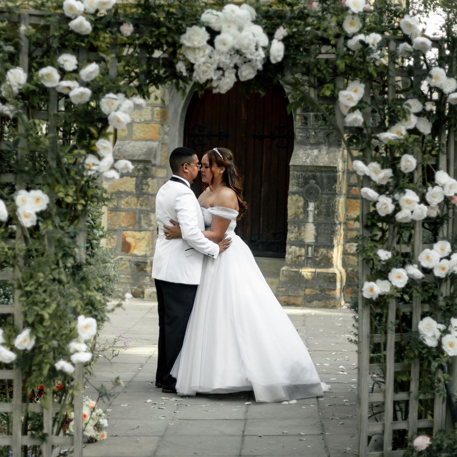 Bride and groom kissing under a floral arch of white roses at a stone church wedding venue.