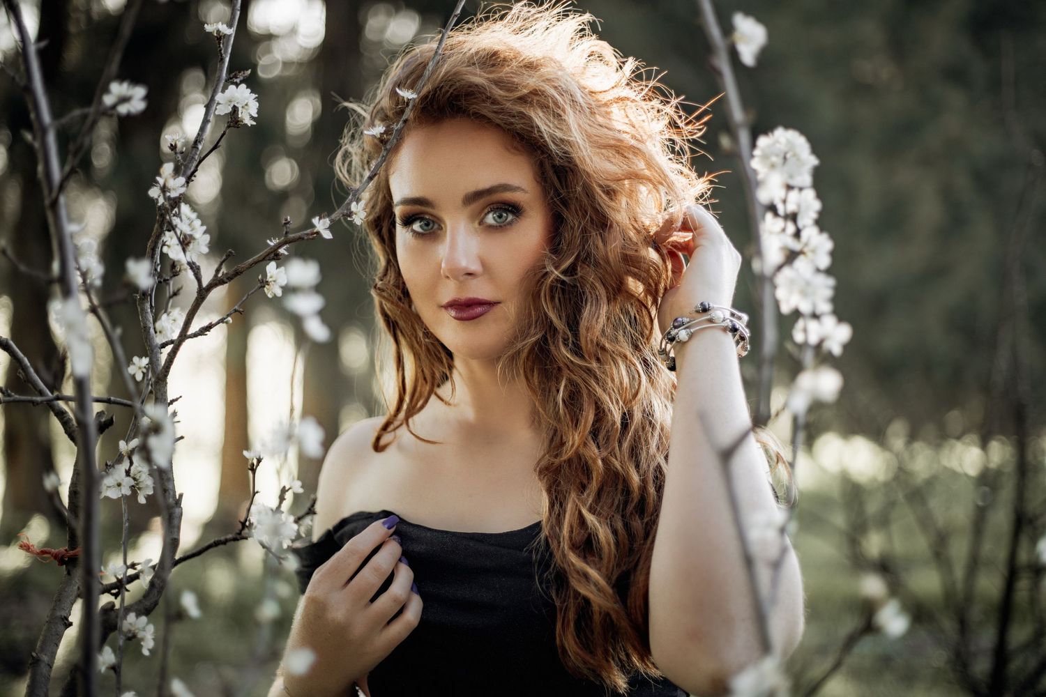 Woman with auburn curls and dark dress stands mysteriously in a forest setting with dreamy lighting.