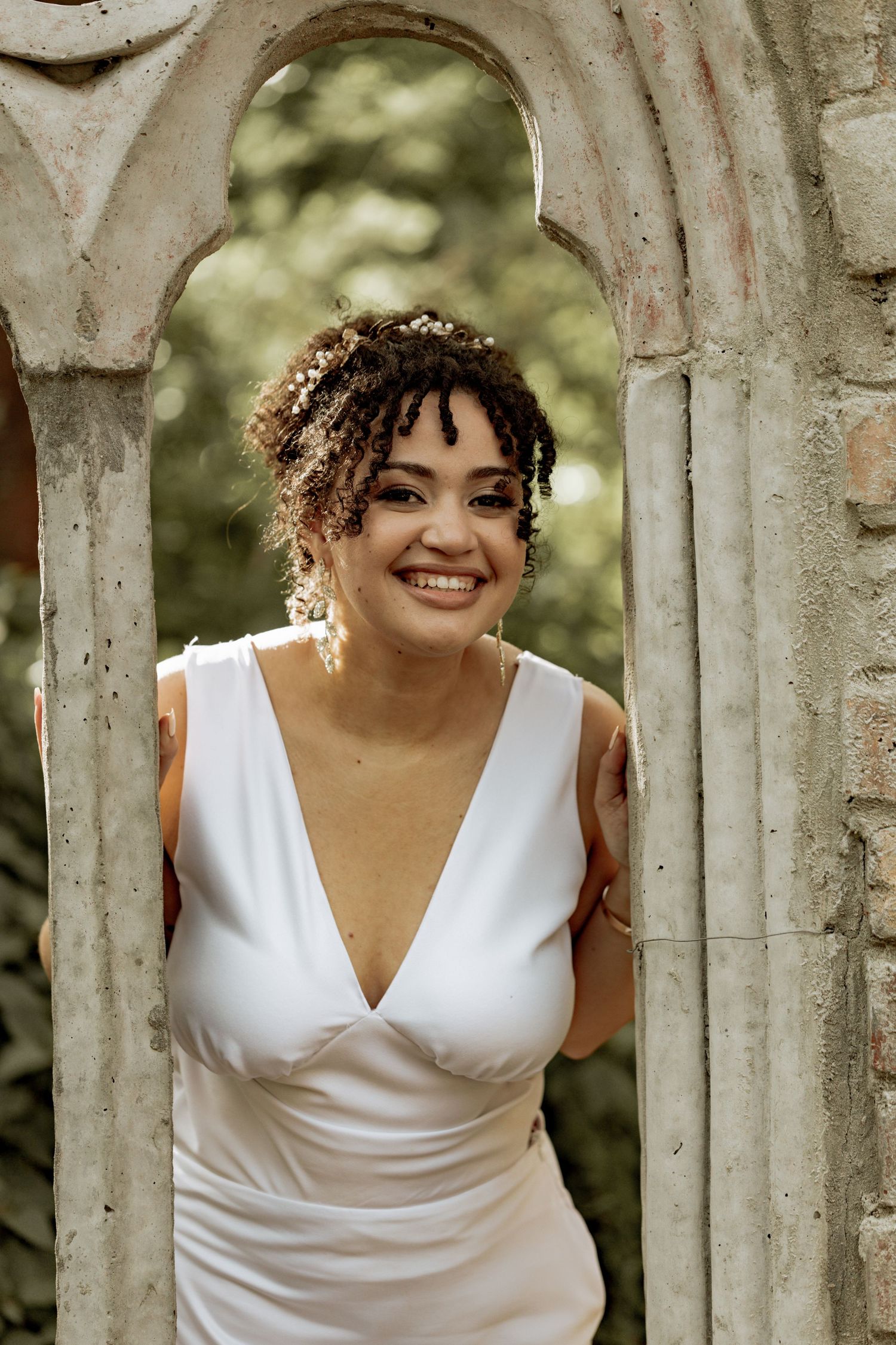 A woman in a white V-neck wedding dress poses joyfully between concrete pillars in an outdoor setting.