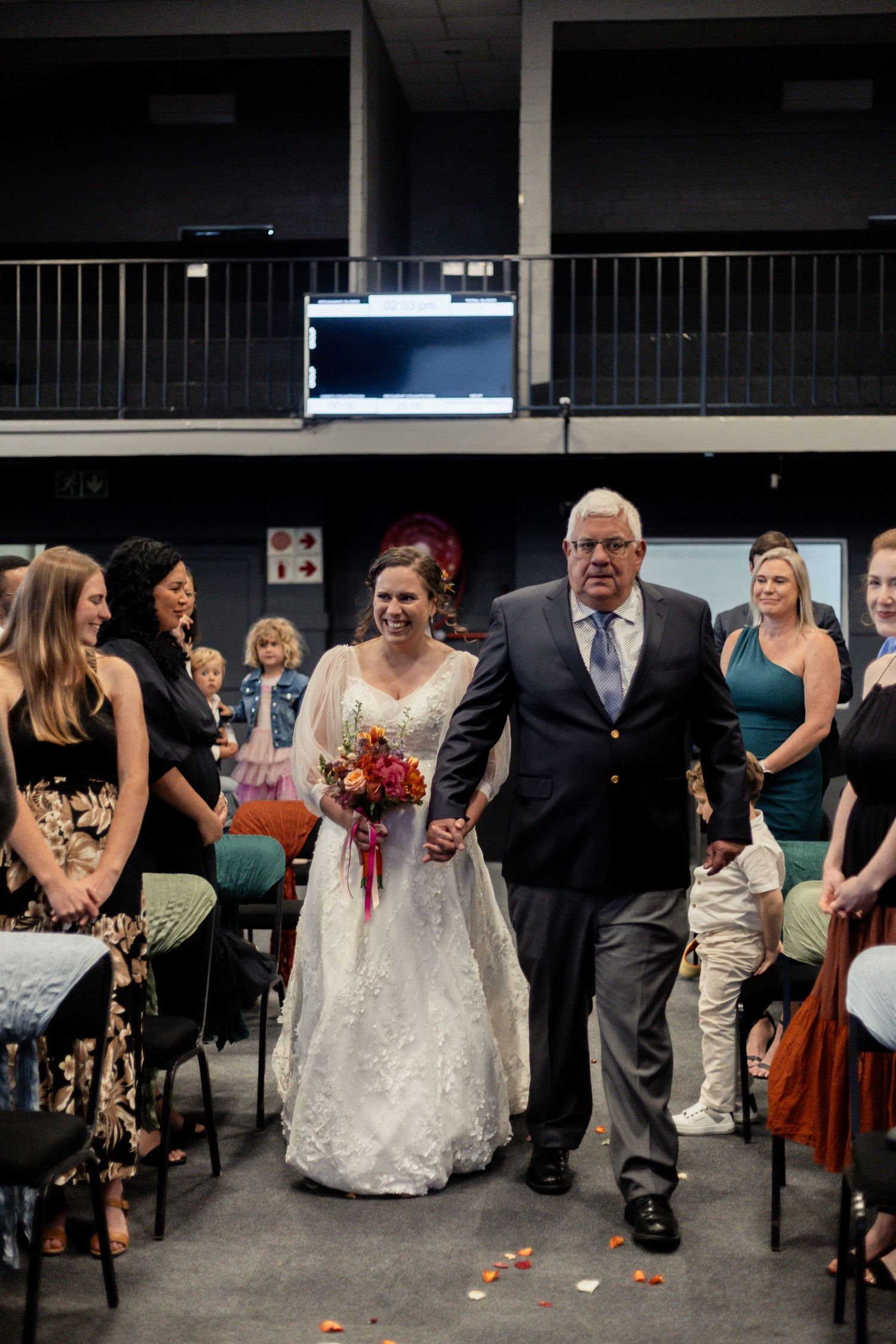 A wedding ceremony processional in a dark indoor venue with scattered rose petals along the aisle.