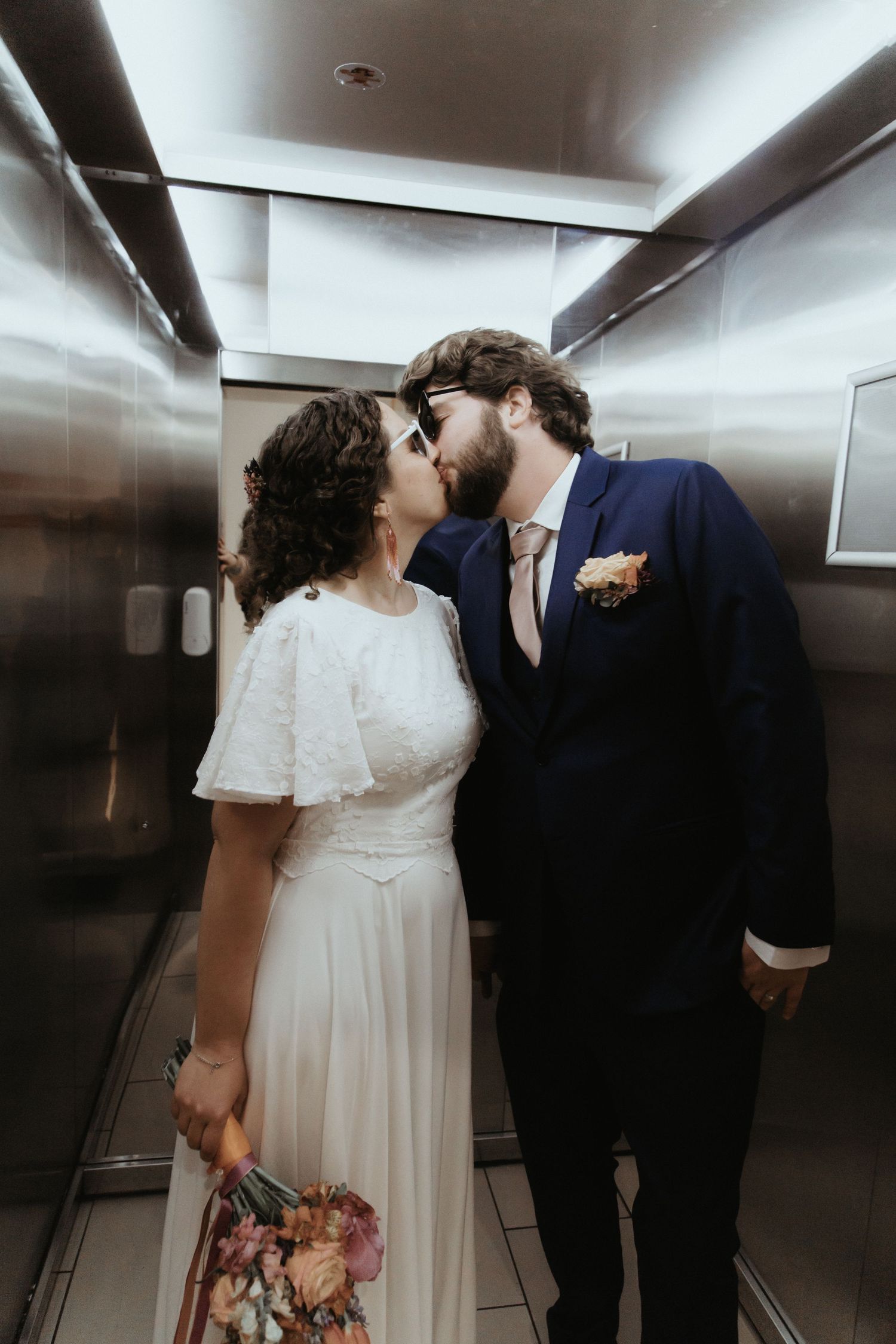 A couple shares an intimate kiss in an elevator while holding a floral bouquet on their wedding day.