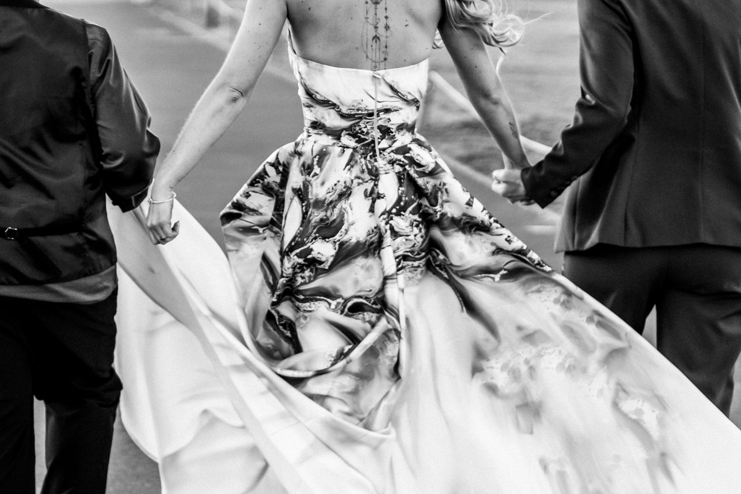 Black and white image showing the flowing skirt of a wedding dress with floral pattern being held by people on either side.