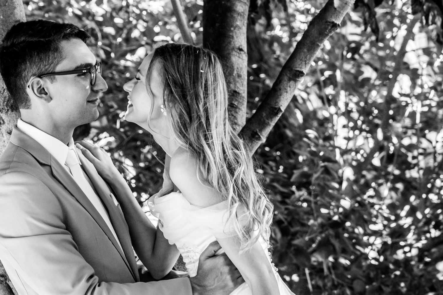 Black and white romantic portrait of a couple embracing under a tree in an intimate outdoor moment.
