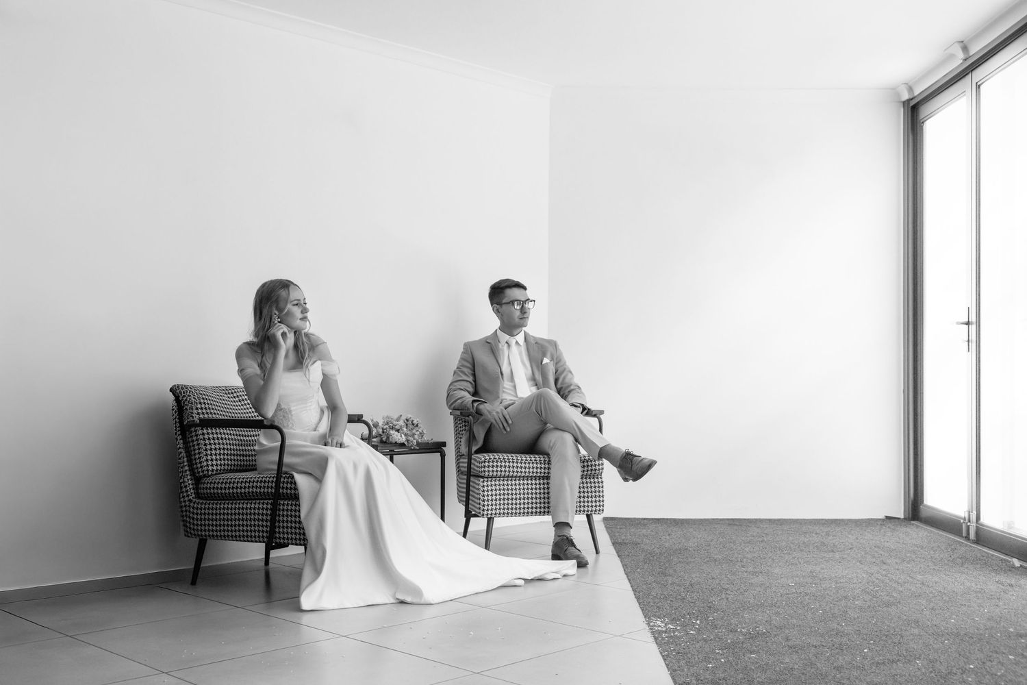Black and white photo of a couple sitting apart in chairs by a window in a minimalist modern space.