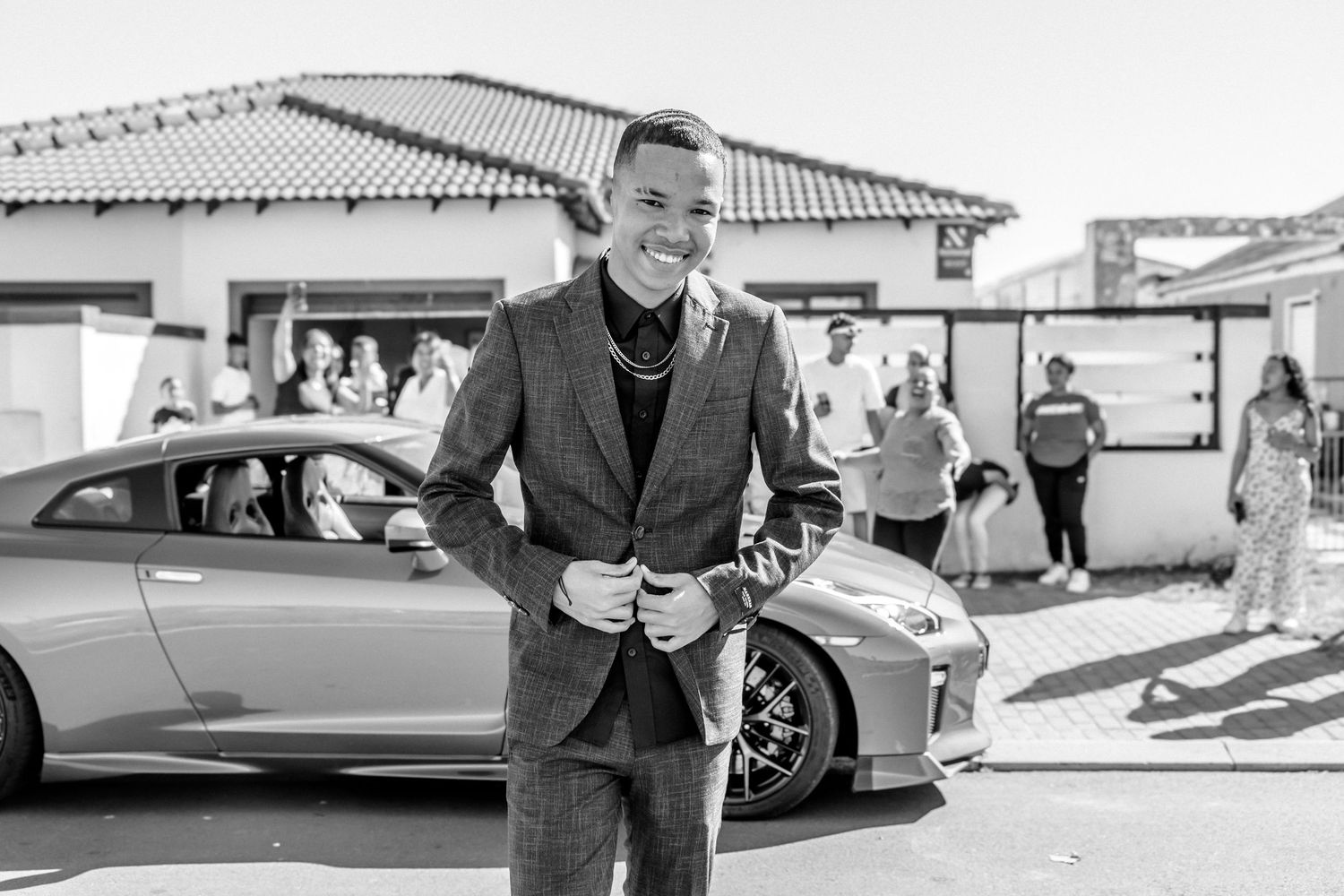 Smiling man in gray suit standing beside luxury sports car outdoors in black and white photo.