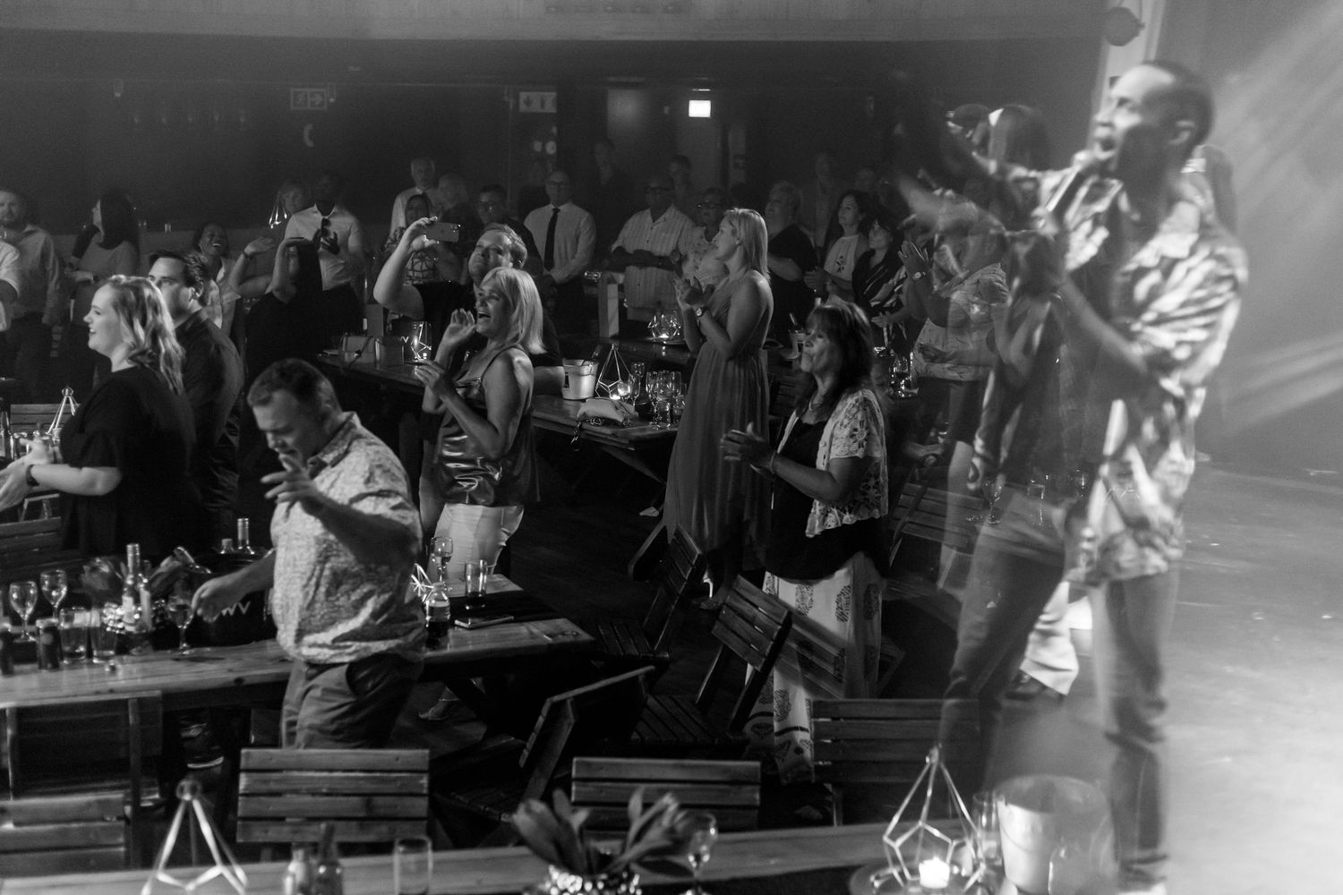 Black and white photo of a lively indoor event with guests mingling and a performer on stage.