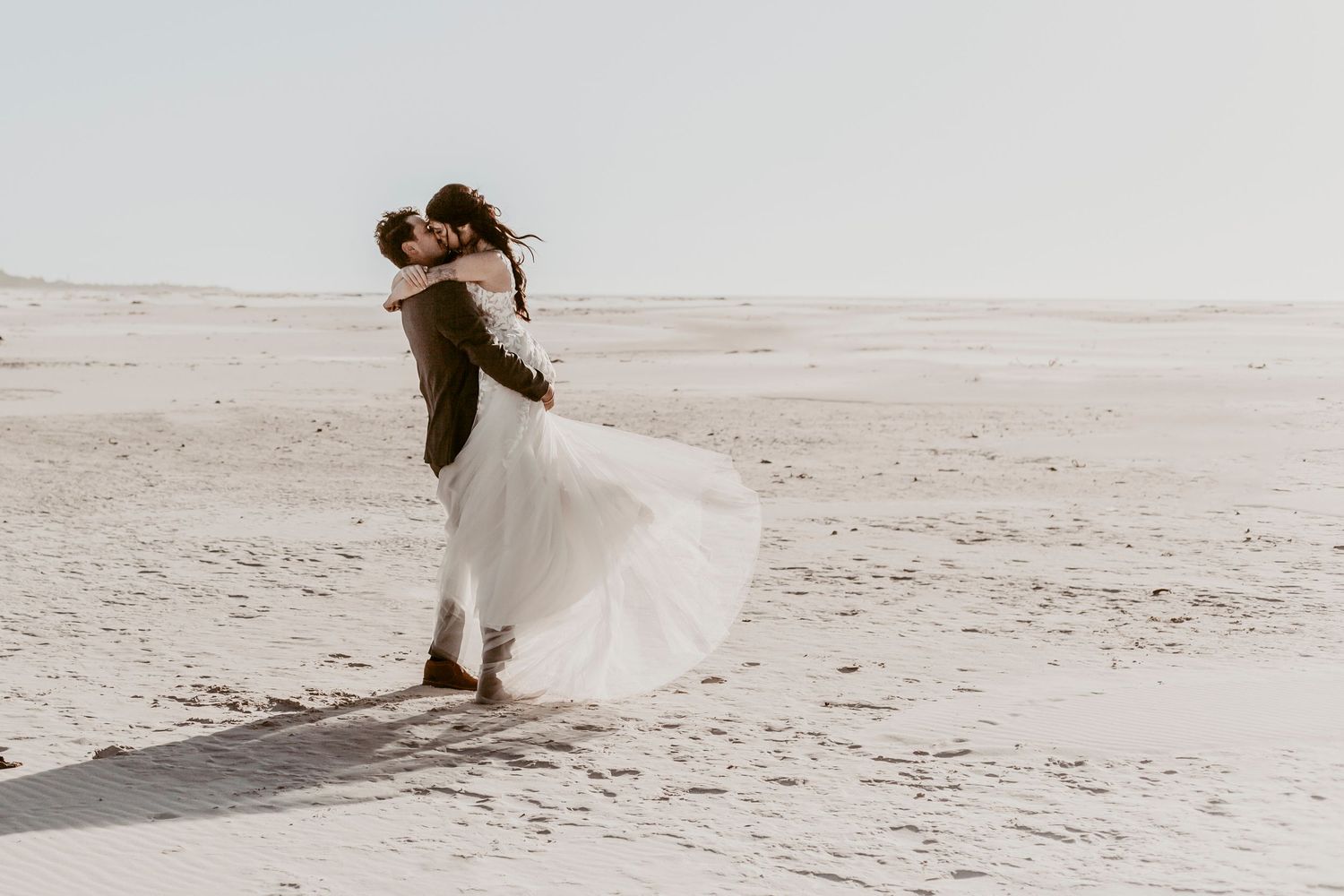 Groom lifting bride in flowing white dress on sandy beach, romantic outdoor wedding photo.