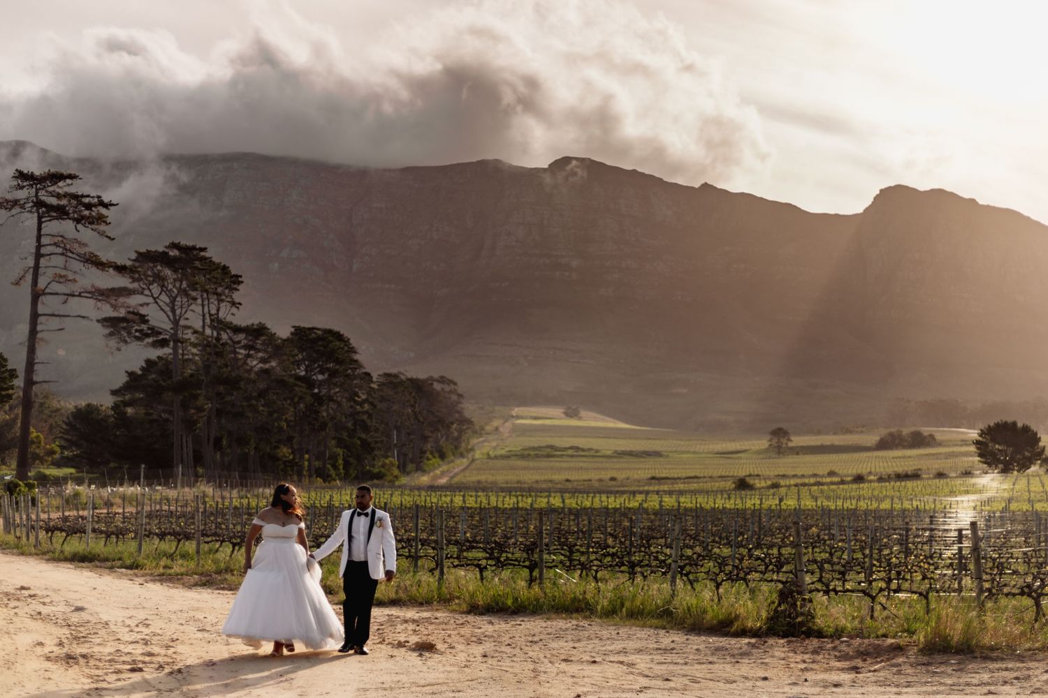 A couple walks along a dirt road beside vineyards with dramatic mountains and sunset in the background in South Africa.