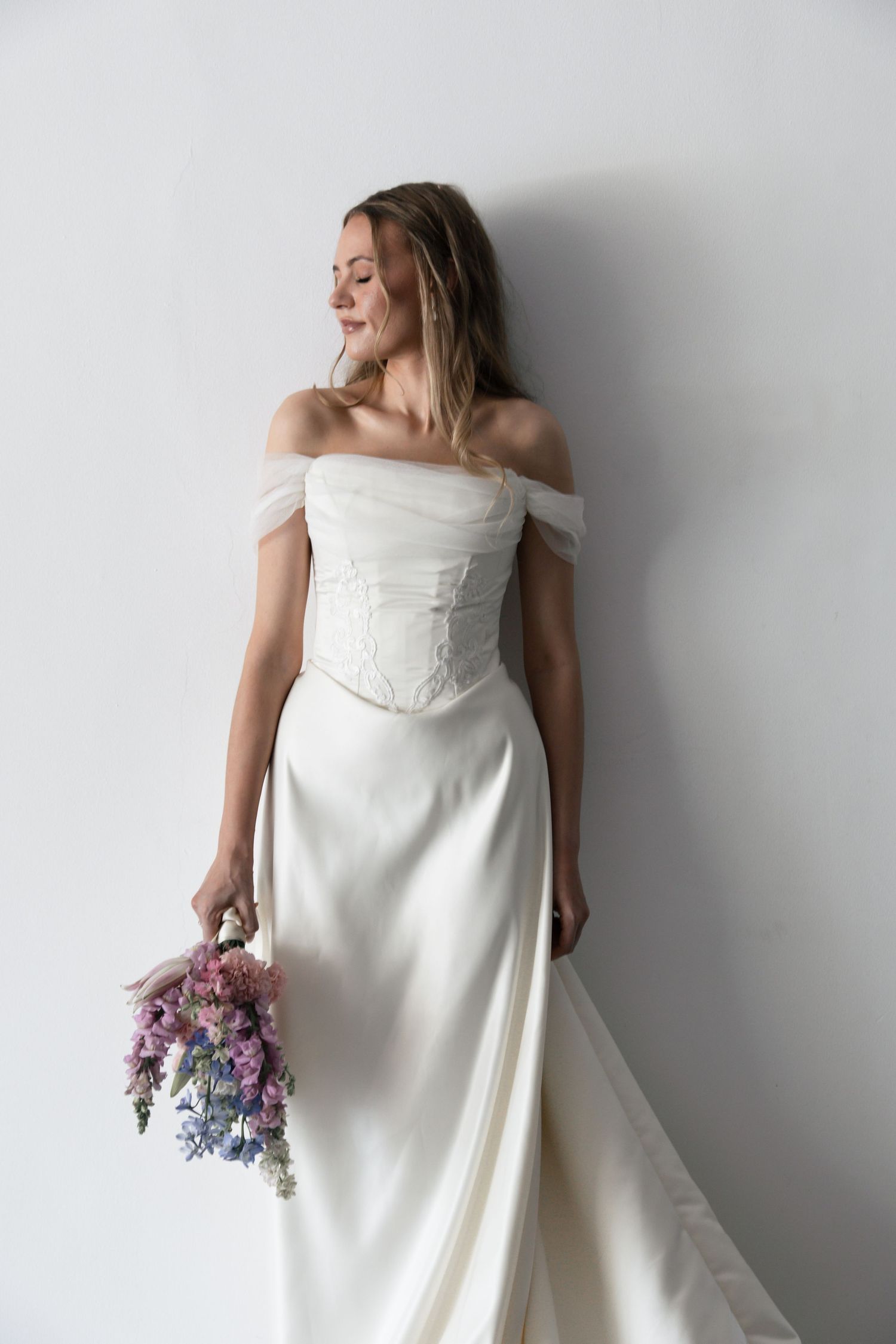 A person in a flowing white wedding dress poses against a white wall while holding a purple and pink floral bouquet.