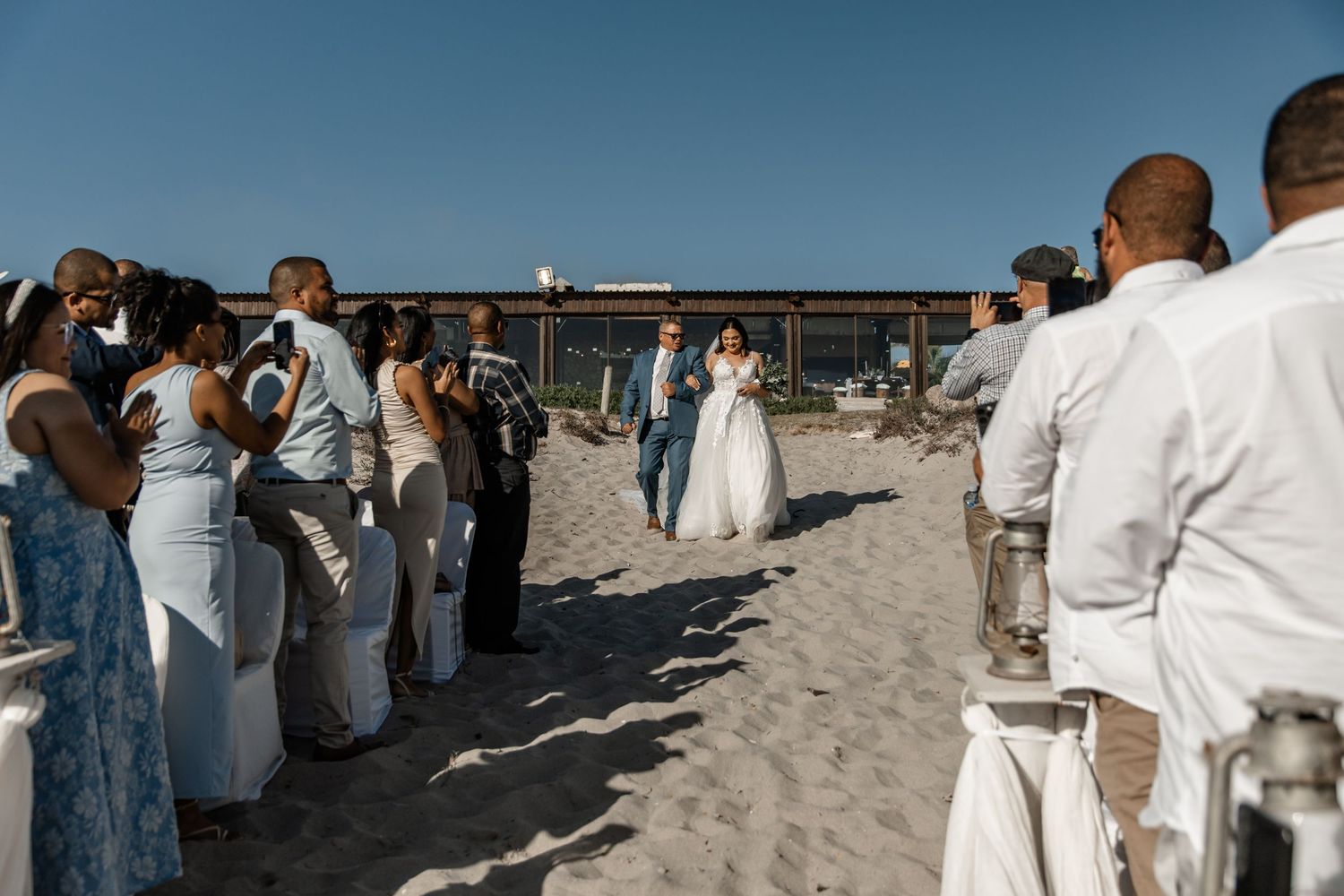 A beach wedding ceremony with guests in white attire gathered on the sand during a sunny day.