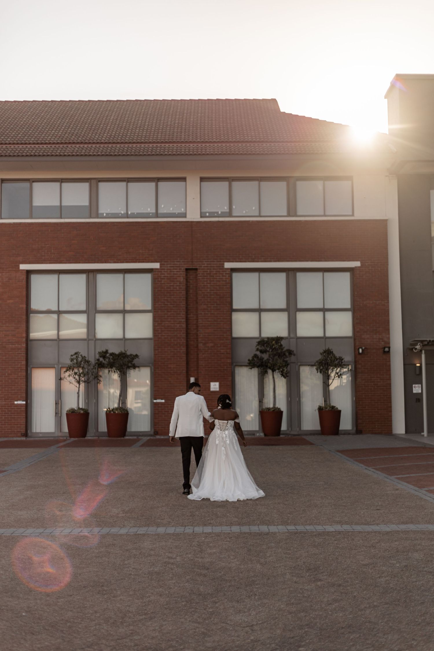 A couple in wedding attire walking outside a modern brick building at sunset.
