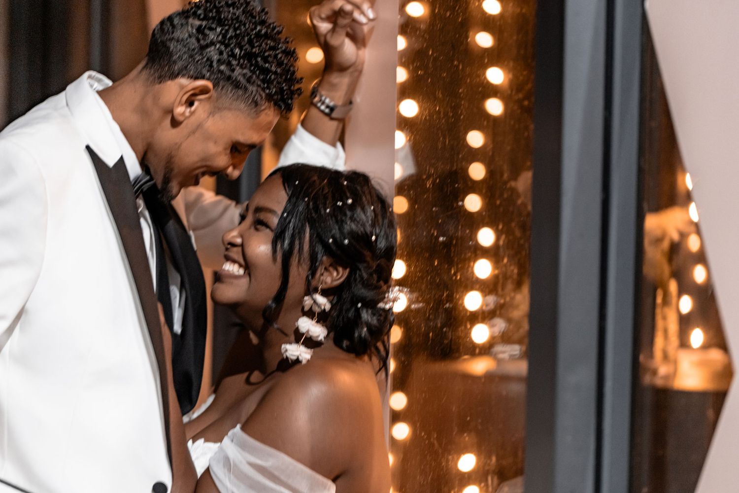 A wedding couple shares a tender, romantic moment together on an outdoor balcony under soft string lights at night.
