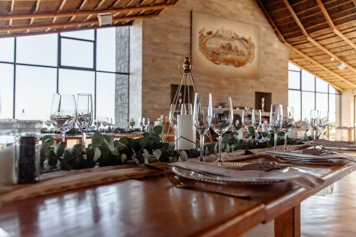 Rustic restaurant dining table with place settings and wine glasses under wooden beamed ceiling with large windows.
