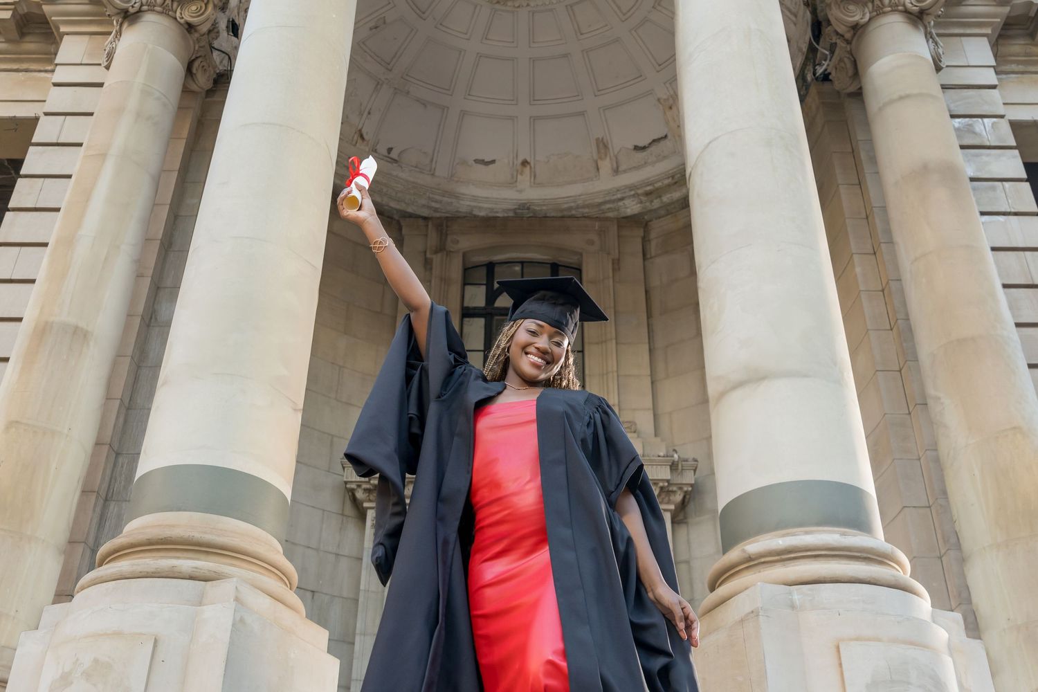 A graduate in cap and gown celebrates with raised arm while standing between classical stone columns.