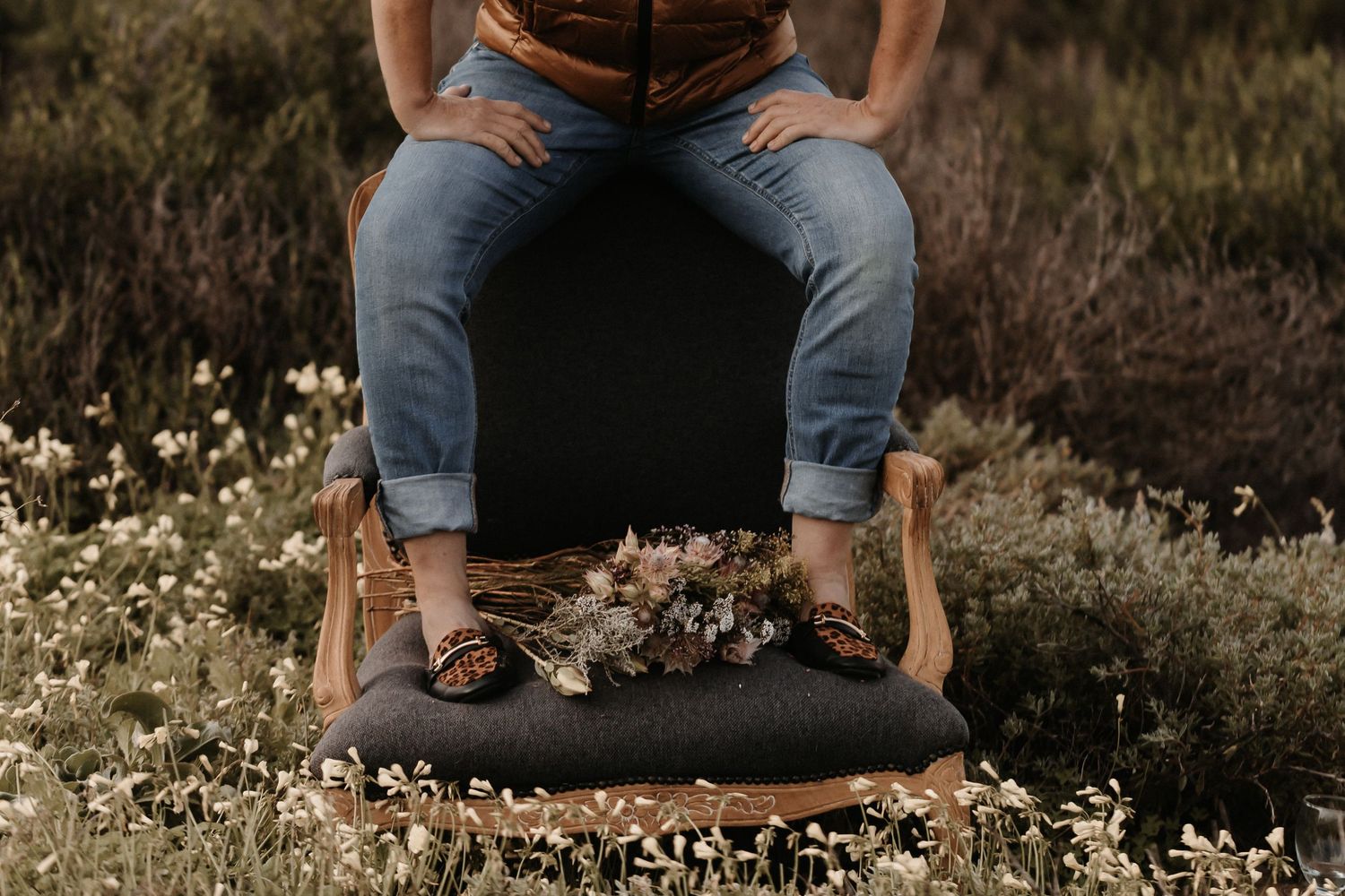 Close up of folded blue jeans and brown leather boots sitting on a black chair outdoors among white wildflowers.
