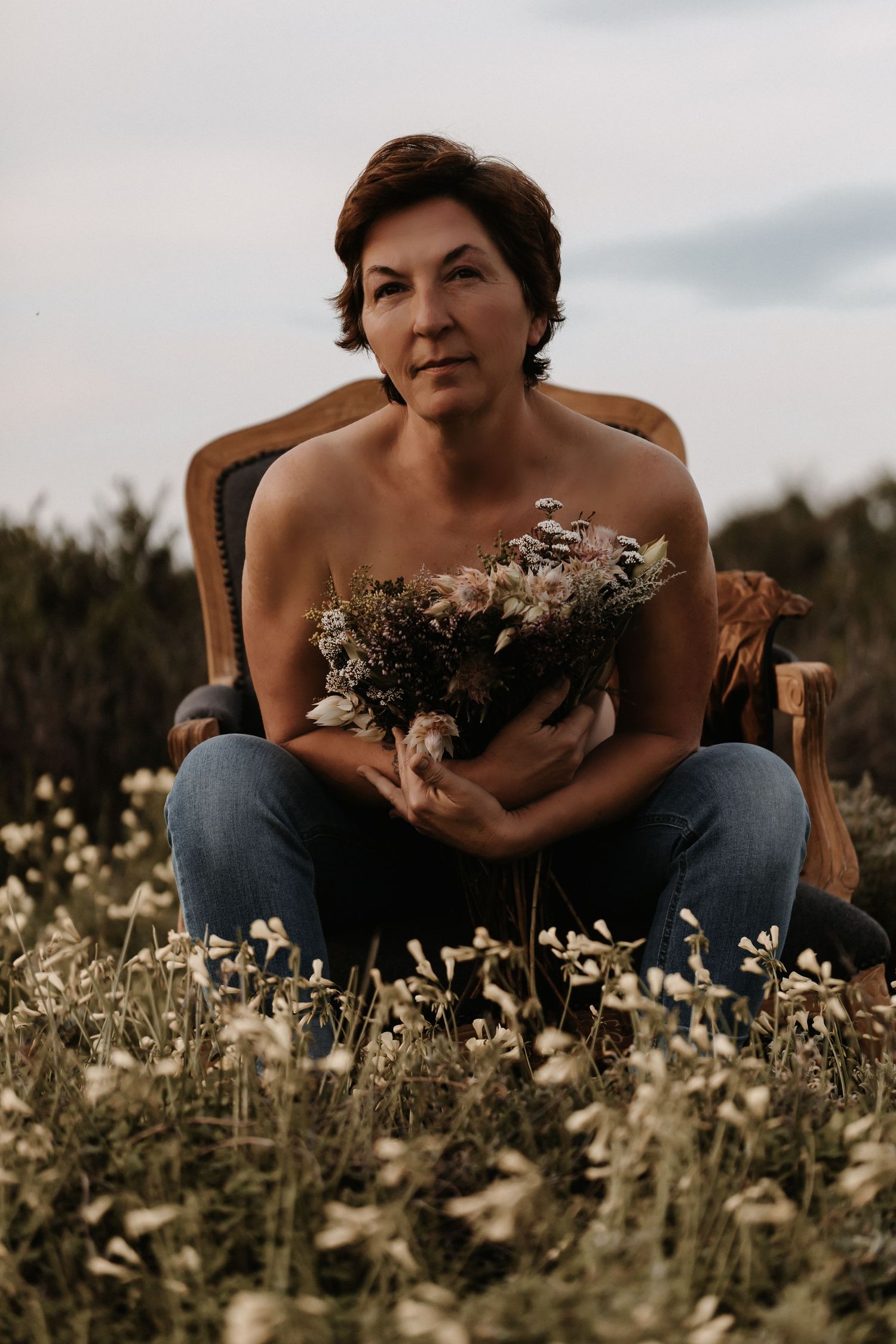 A person in jeans sits in a vintage chair amid a field of wildflowers holding a rustic dried flower bouquet.