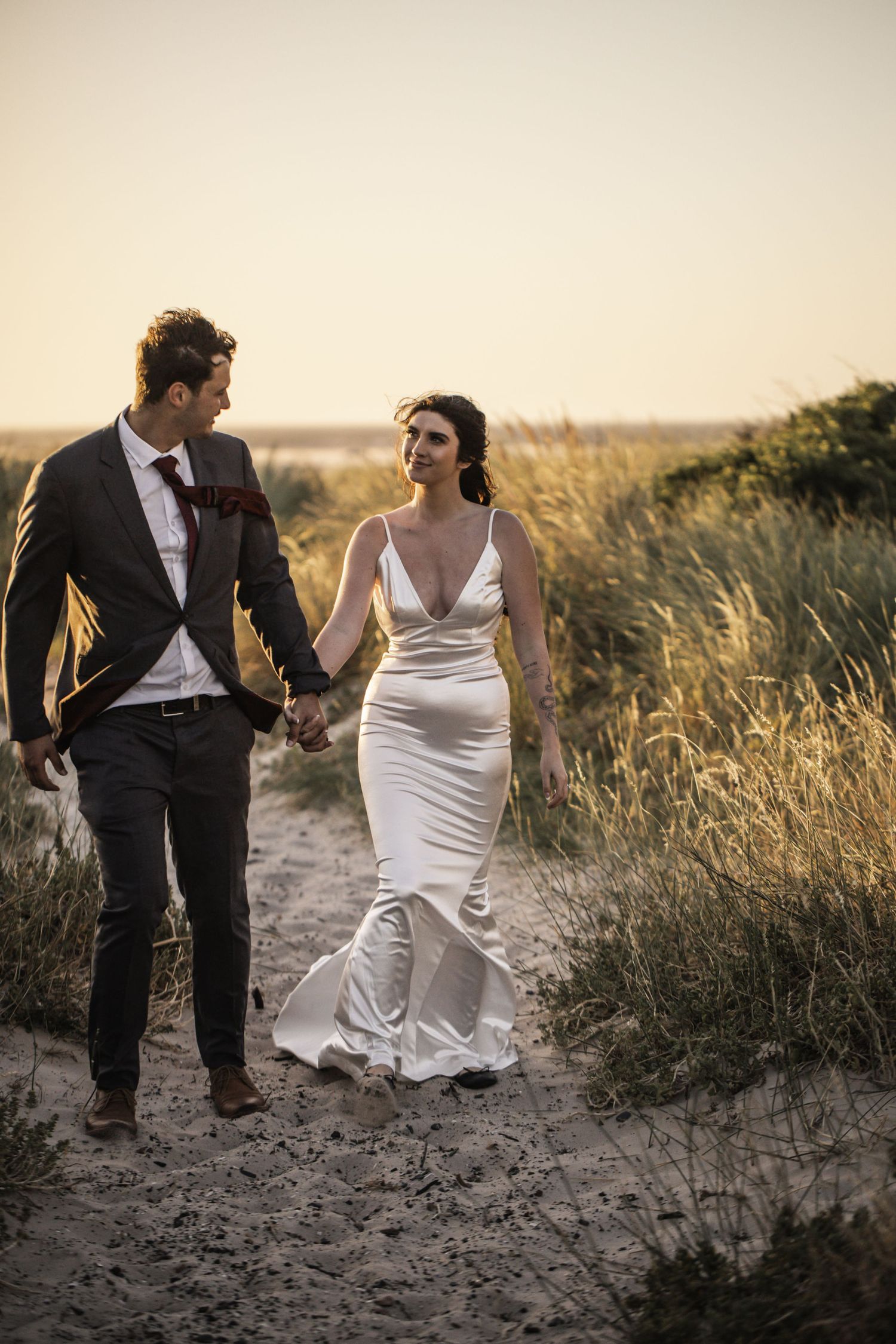 Bride and groom walk hand-in-hand along a sandy beach path at sunset, surrounded by tall golden grasses.