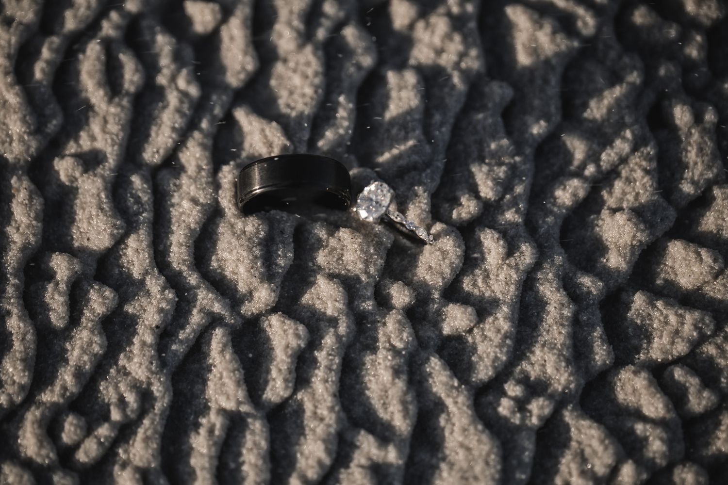 Two rings, a black band and diamond engagement ring, resting on textured sandy ground with dramatic shadows.