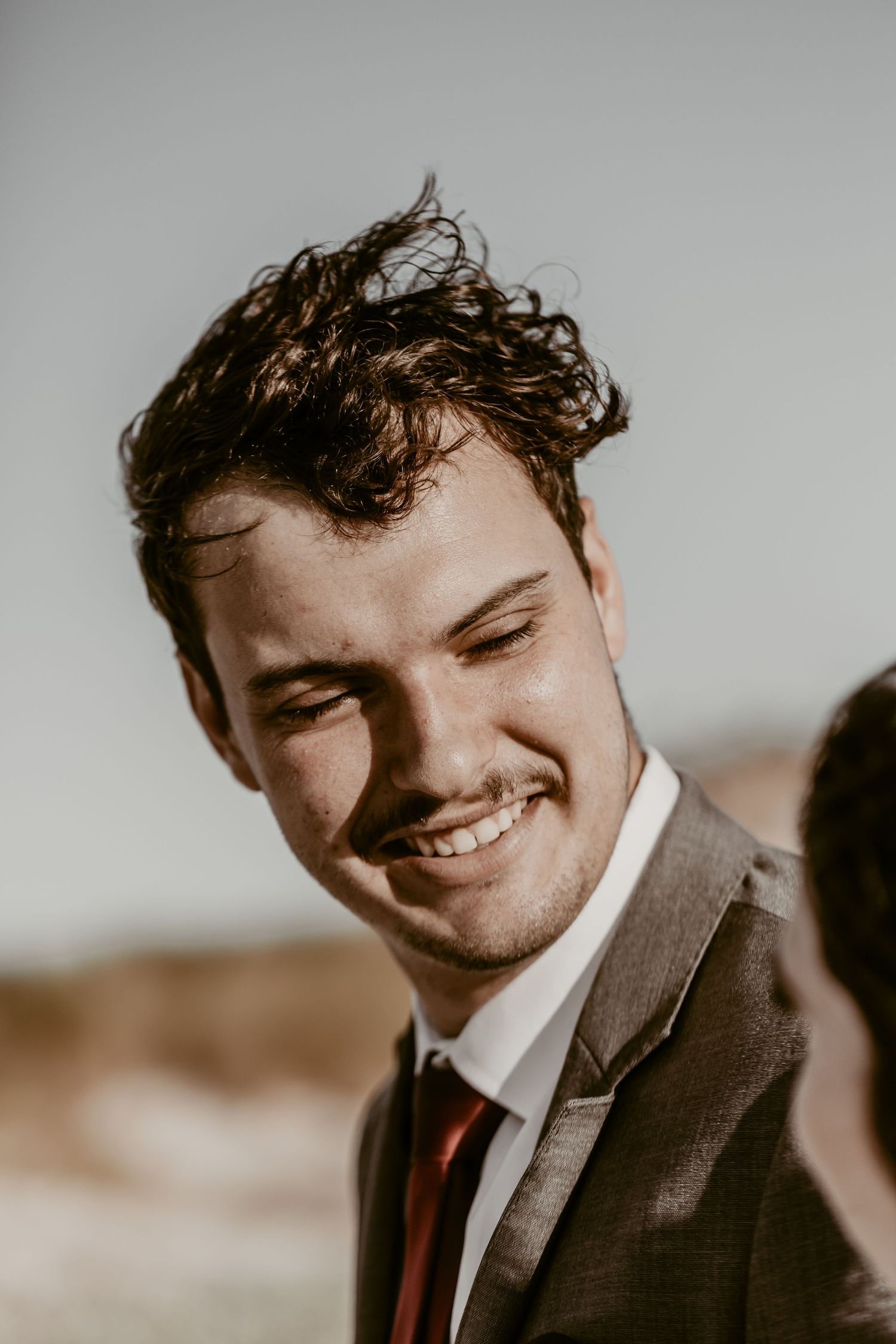 A smiling groom in a gray suit and red tie with curly hair poses outdoors on a bright sunny day.
