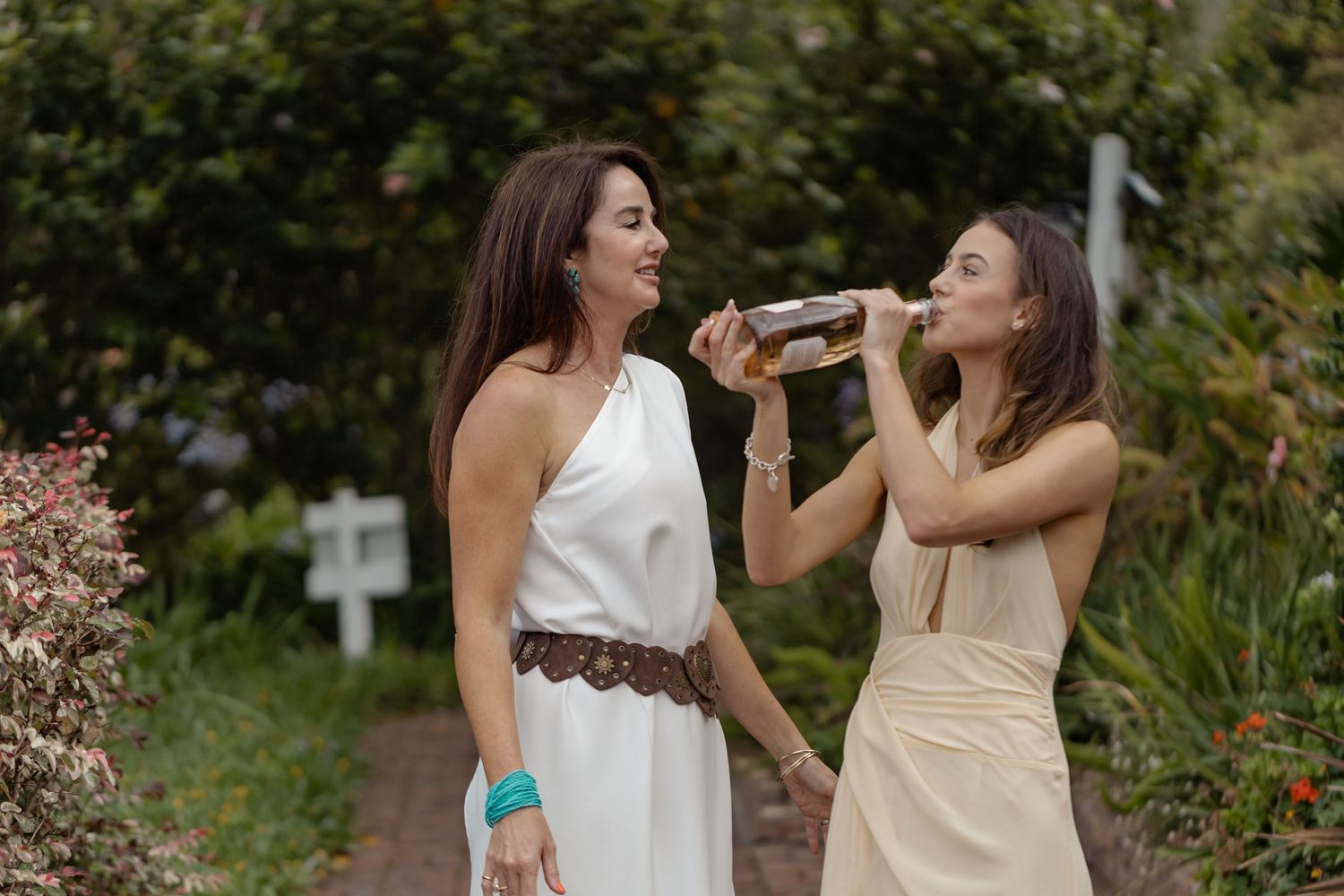 Two women in elegant dresses share a bottle of wine outdoors in a lush garden setting.