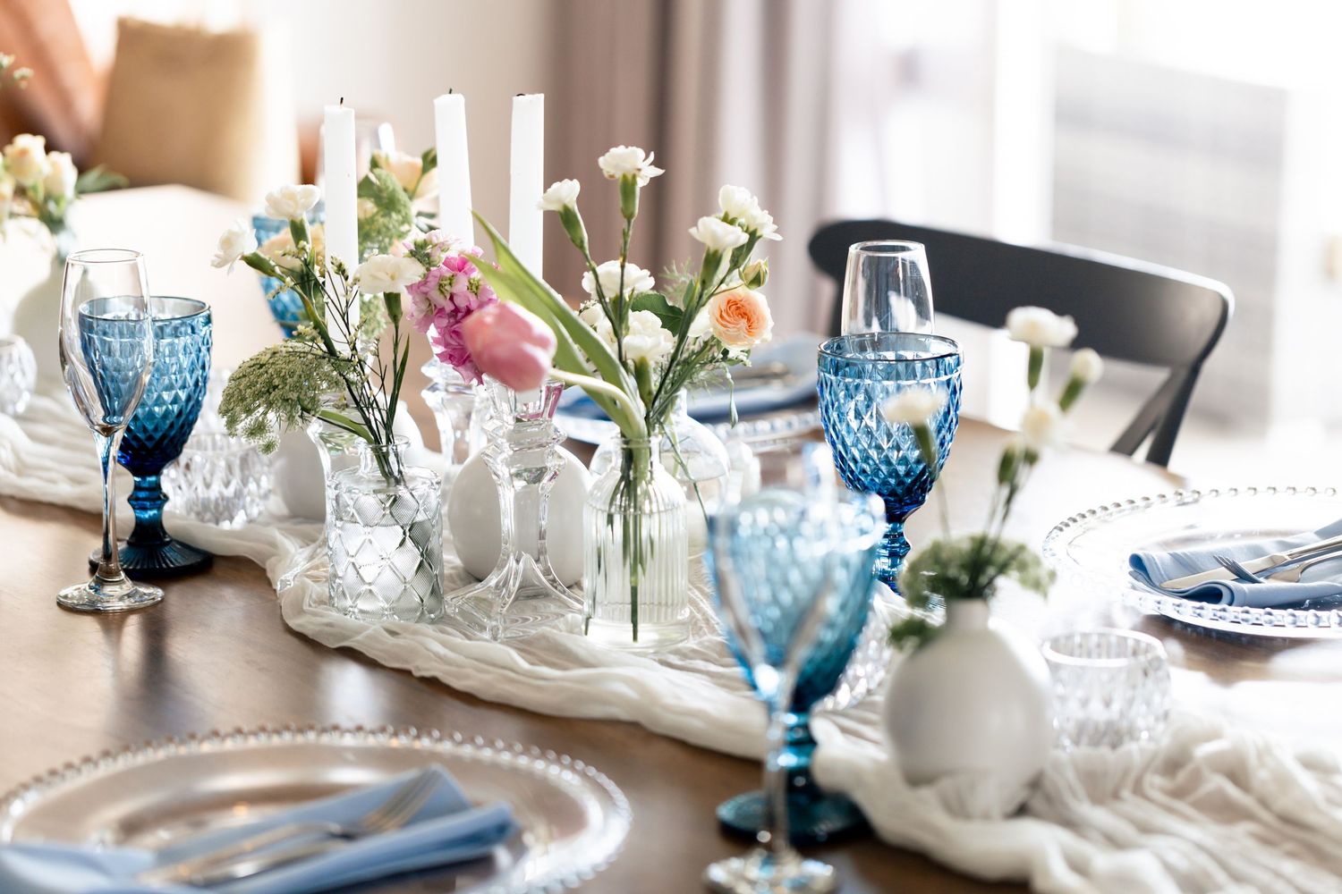 Elegant table setting with blue vintage goblets, white flowers in vases, and natural light streaming through windows.