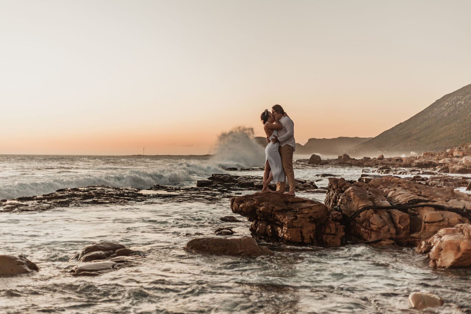 A couple stands on rocky coastal cliffs during golden hour sunset with ocean waves crashing against the shore.