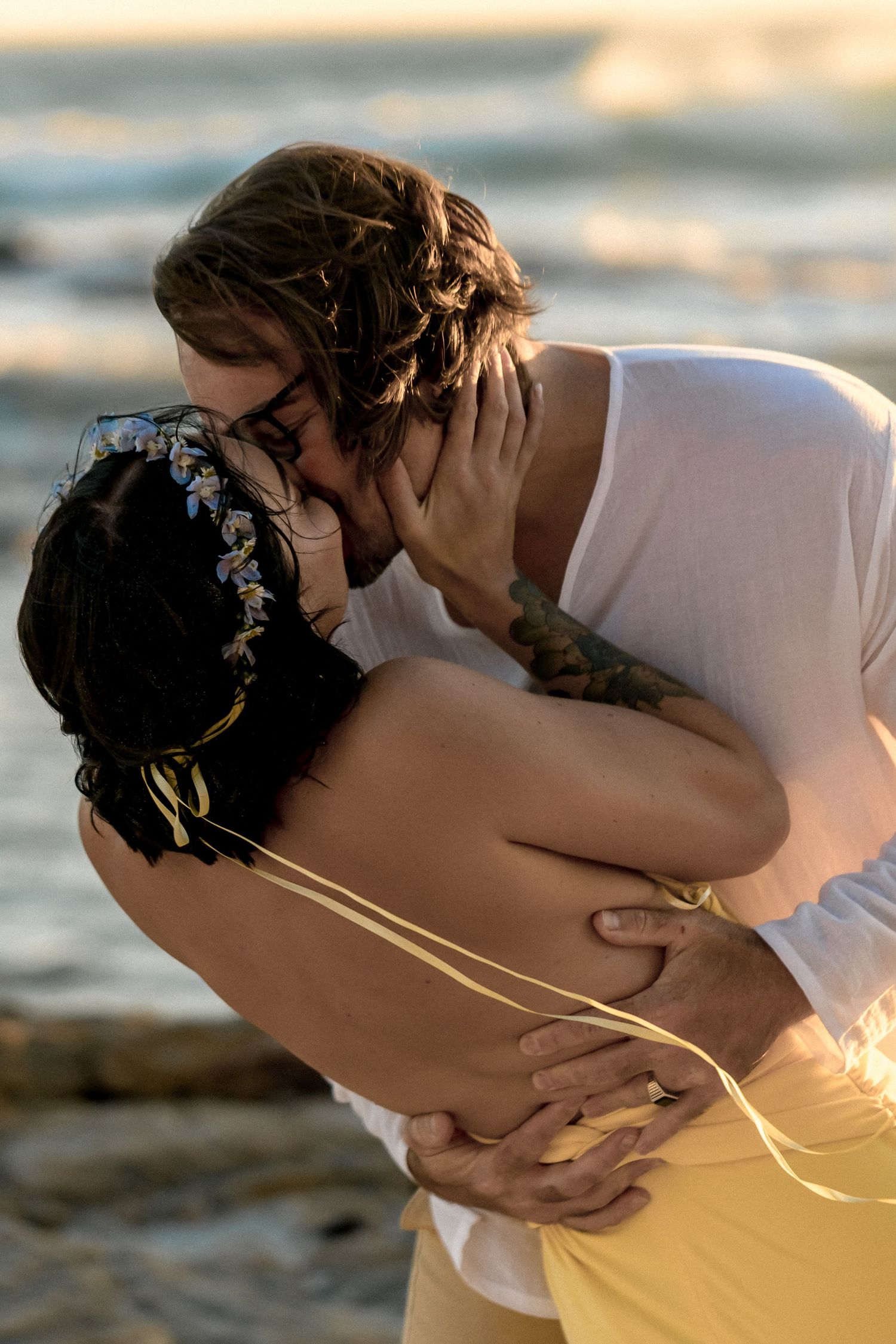 A romantic beachside wedding couple shares an intimate kiss at sunset, with gentle waves in the background.