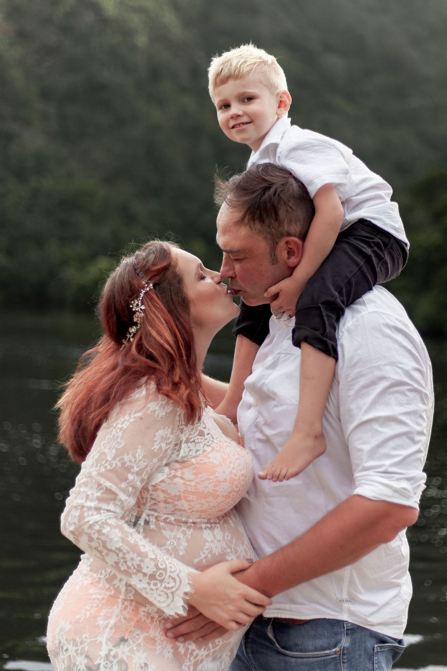 A family shares a tender moment outdoors near a lake, with a pregnant woman in a lace dress and child sitting on shoulders.