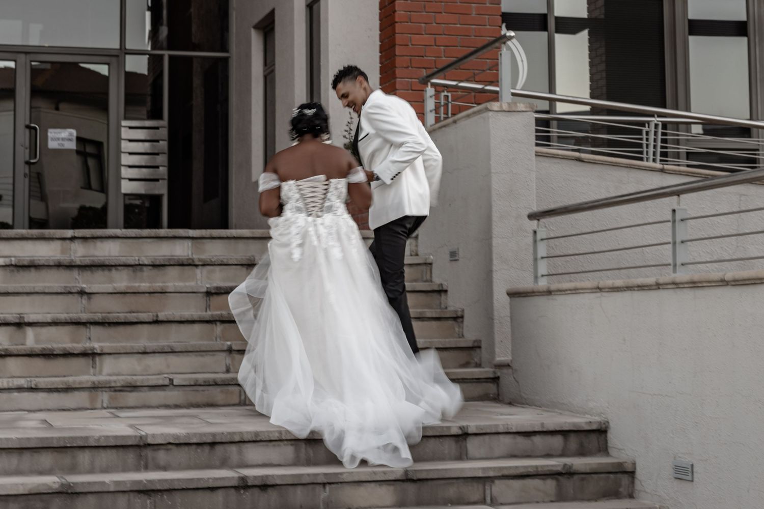 A person in an elegant white wedding dress poses on stone steps by a brick building exterior.