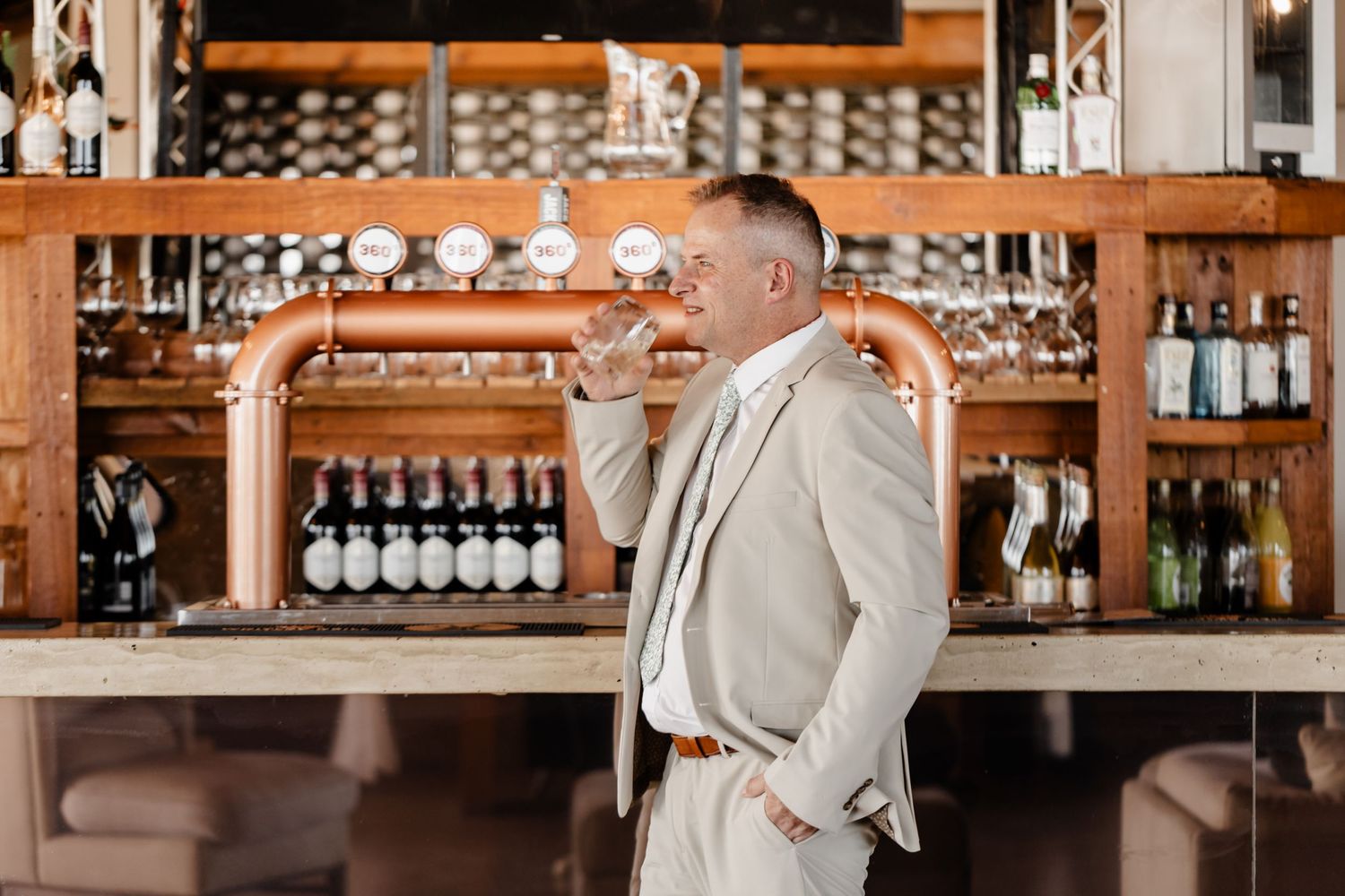 Professional dressed in a beige suit standing at an elegant bar counter with wine bottles and glassware on display.