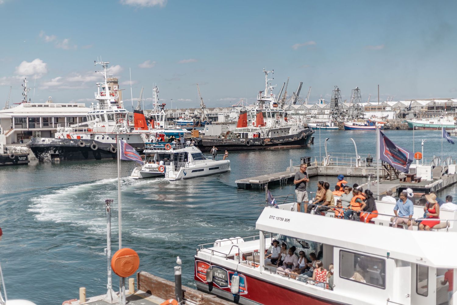 Tourist boats and fishing vessels docked at a busy harbor with cranes and maritime infrastructure in the background.