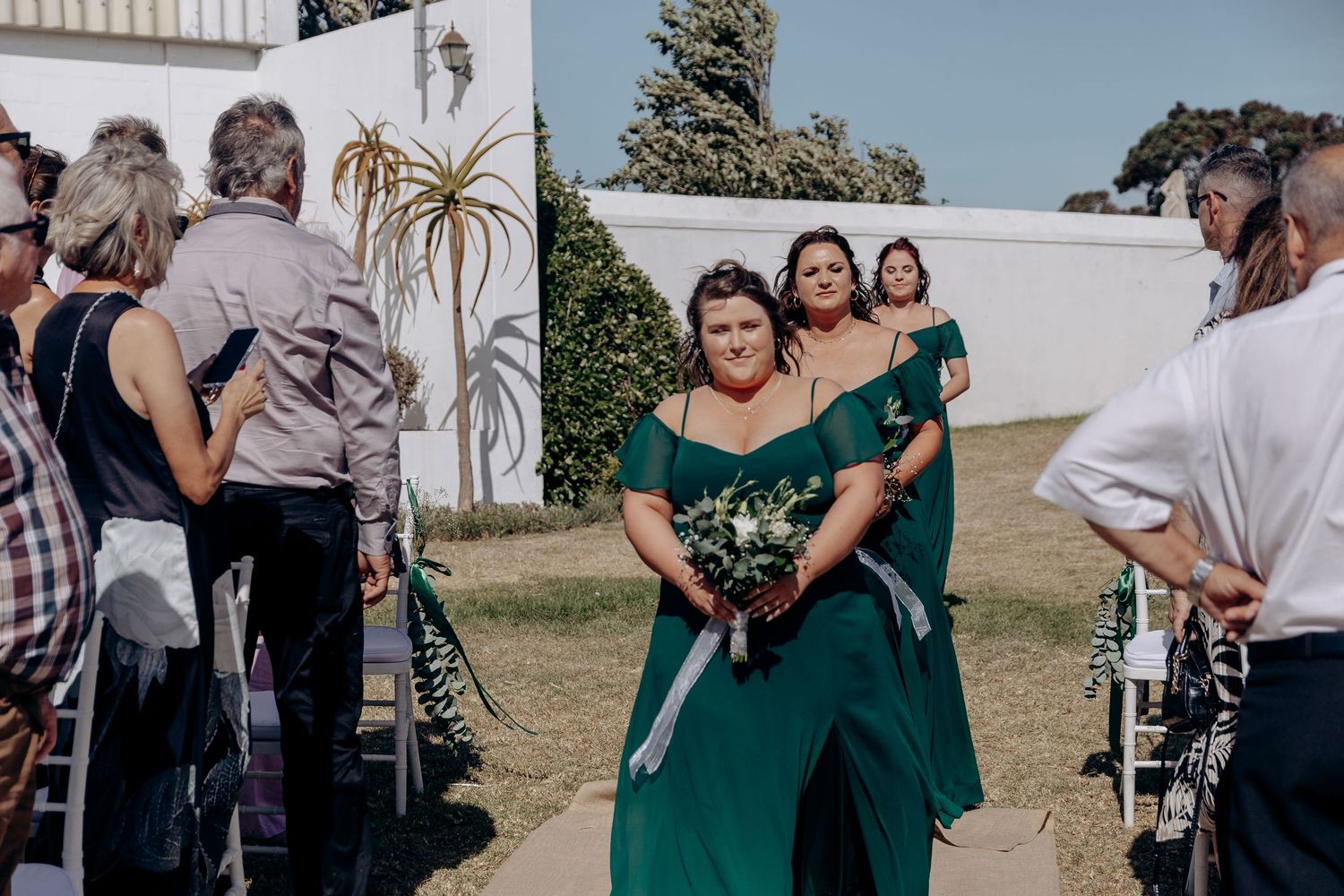 Bridesmaids in emerald green off-shoulder dresses walk down outdoor wedding aisle carrying bouquets.