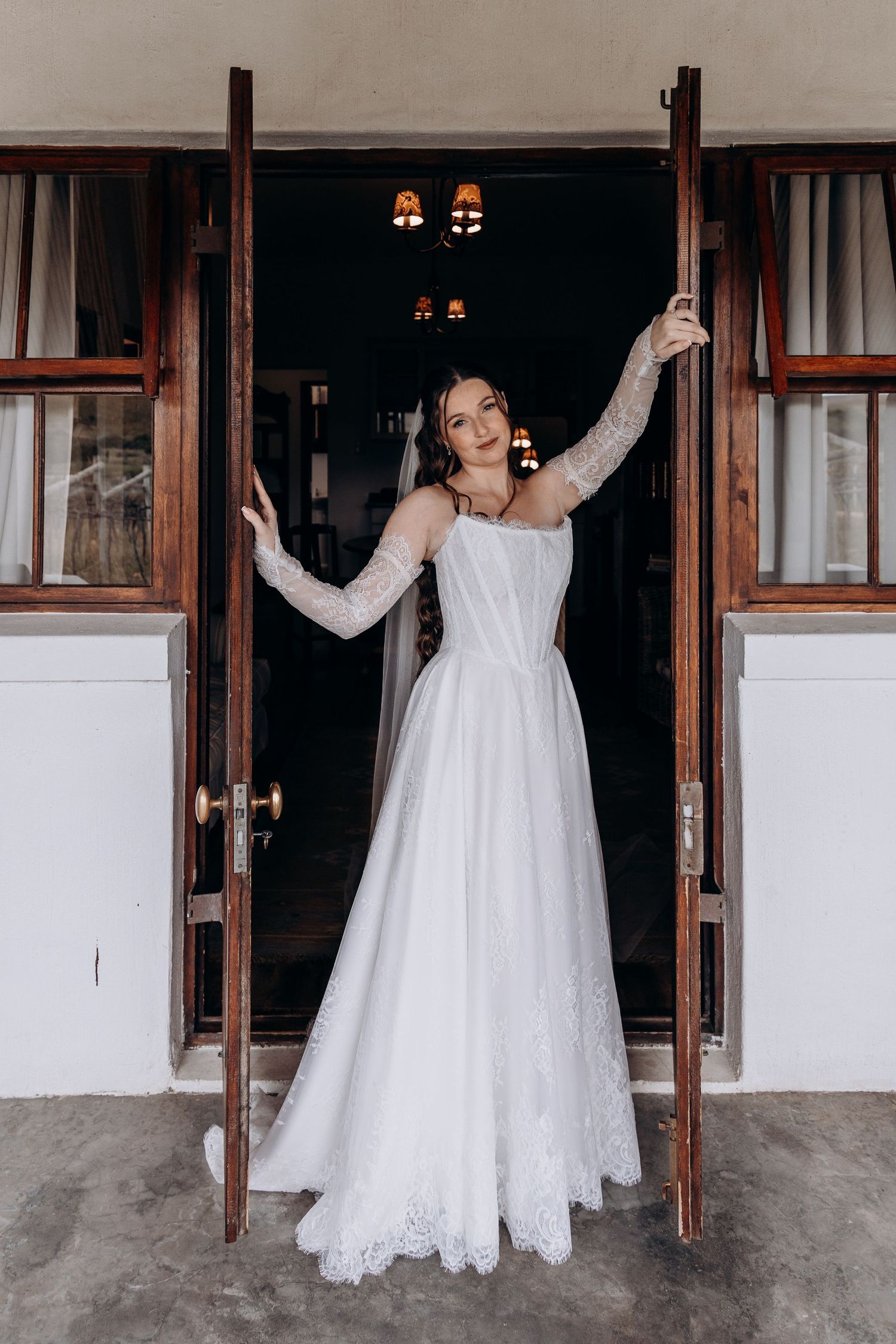Bride in white lace long-sleeve wedding gown stands in open wooden doors, arms raised, smiling elegantly.