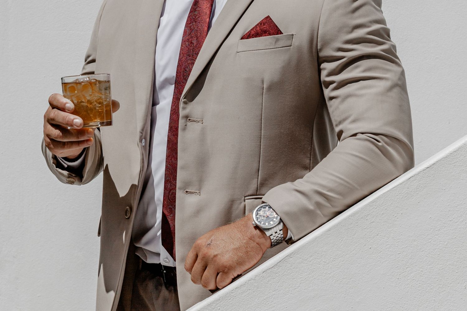 Professional wearing a beige suit and red tie holds a glass of whiskey while standing against a white background.
