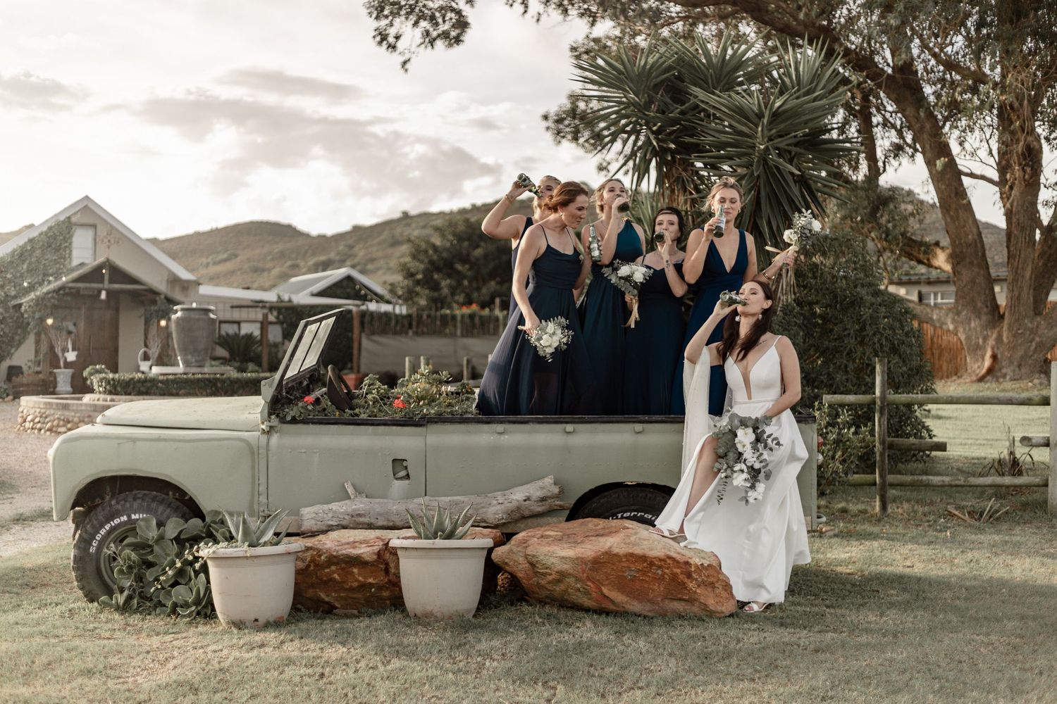 Wedding party poses with vintage Land Rover against mountain backdrop at sunset.