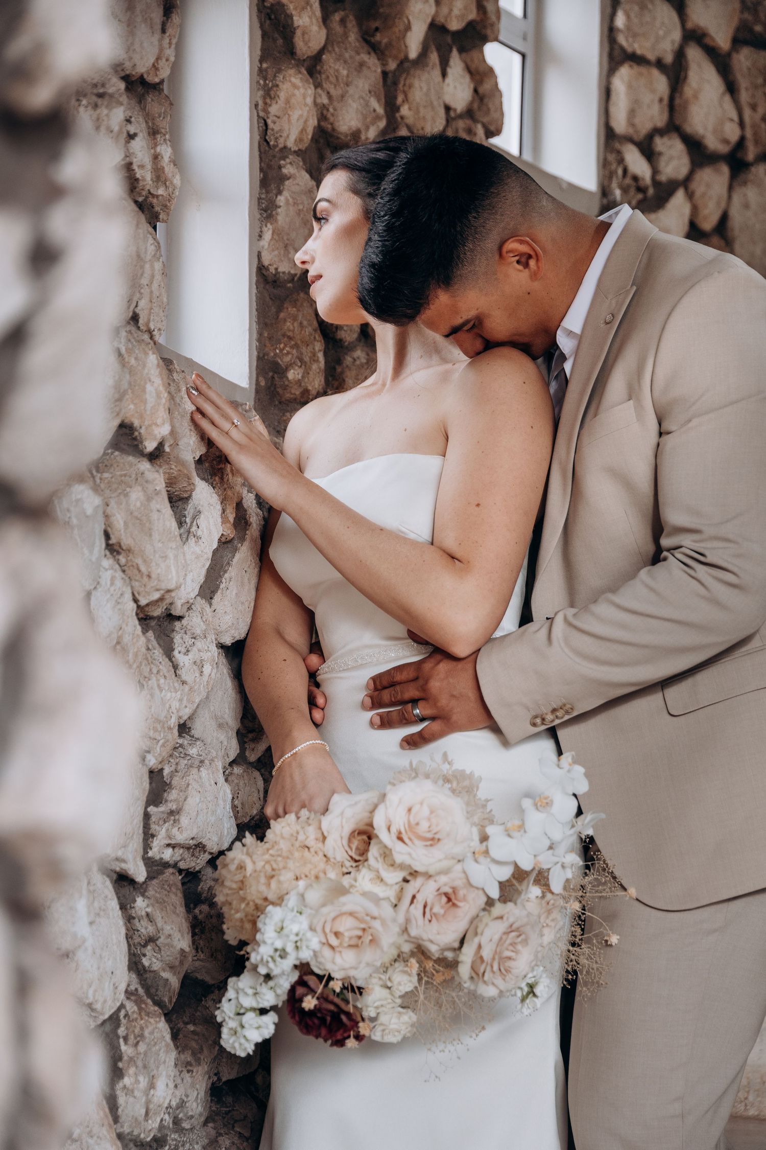 Groom in beige suit kissing bride in white strapless gown against stone wall, holding lush ivory and blush bouquet.