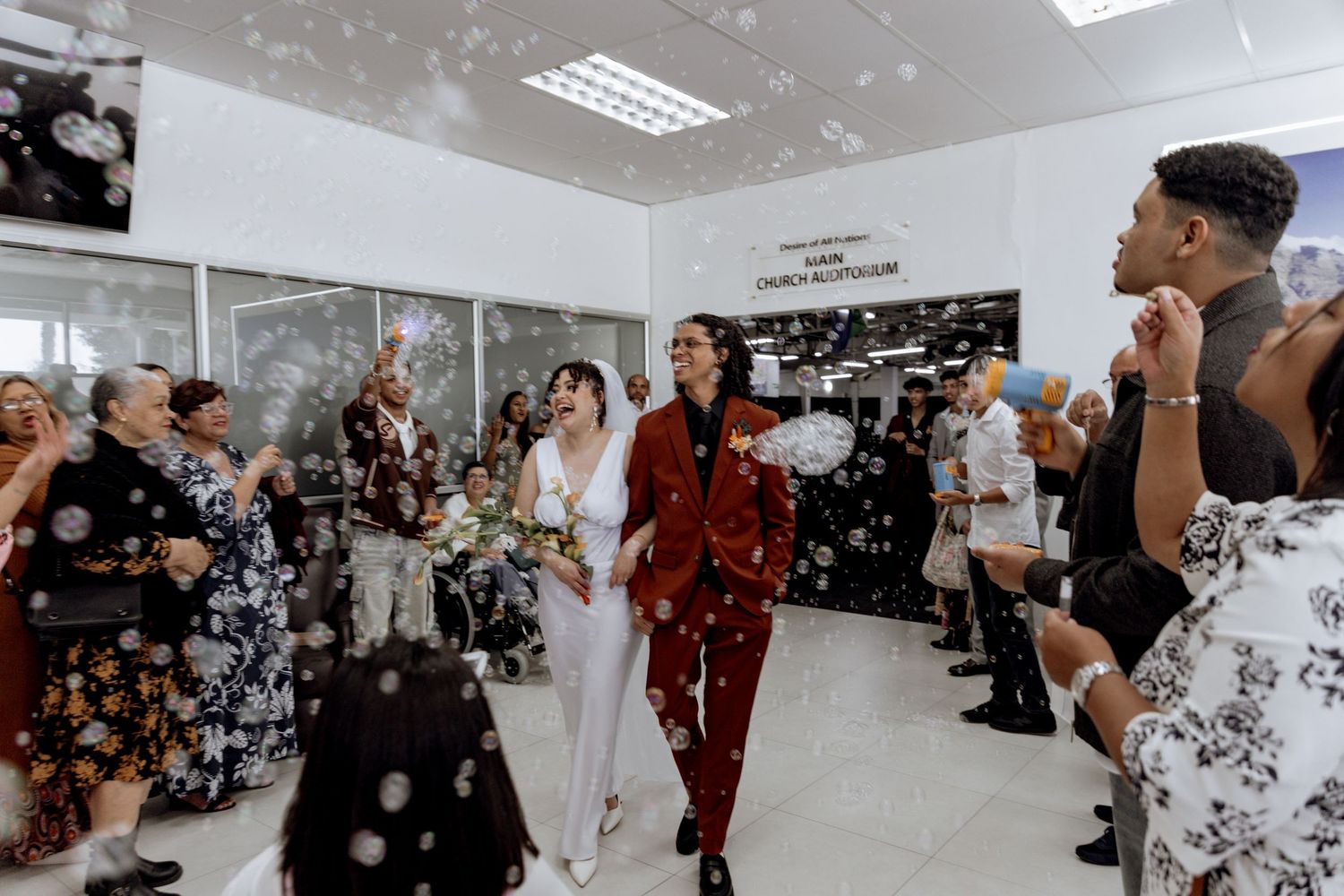 Wedding party celebrates in a white room with bubbles floating through the air and people in formal attire lining both sides.