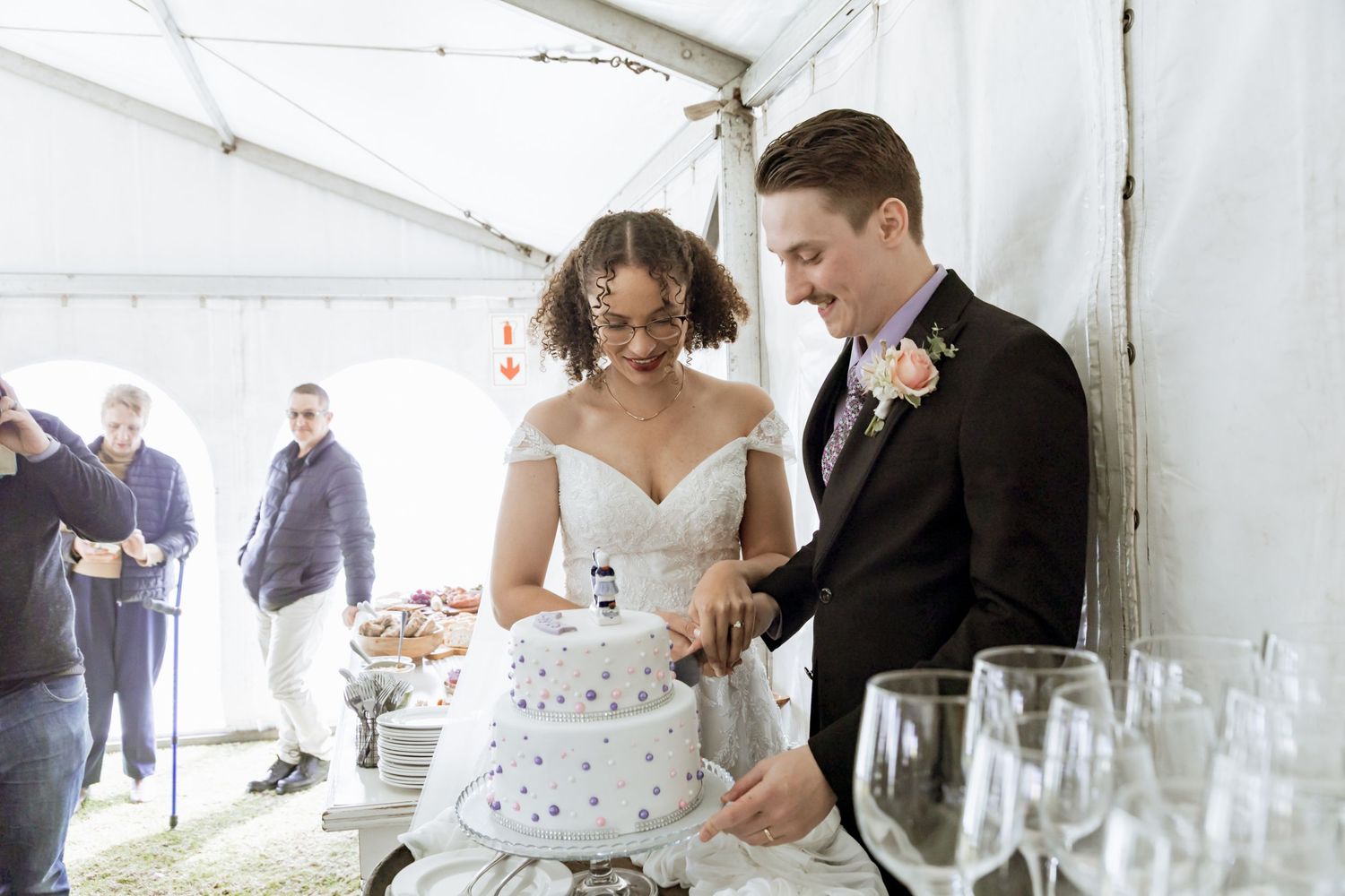 Newlyweds slice into their white tiered wedding cake at an elegant reception under a white tent.