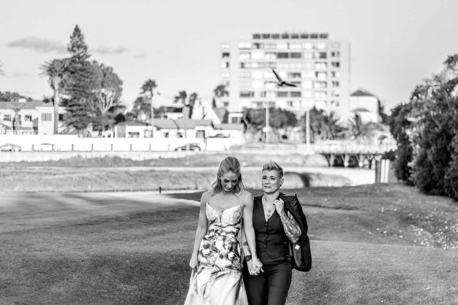 Black and white photo of a couple walking together outdoors with buildings and palm trees in the background.