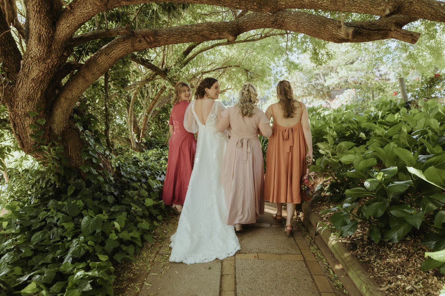 A group in formal attire walks along a lush garden path beneath towering trees and greenery.