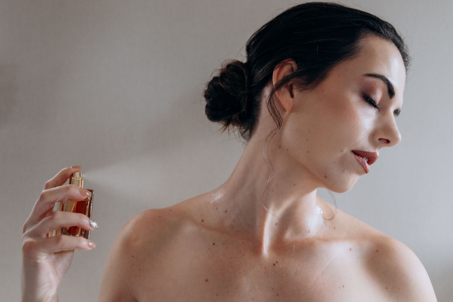 Woman with dark hair spraying perfume on her neck, eyes closed, elegant beauty routine shot.