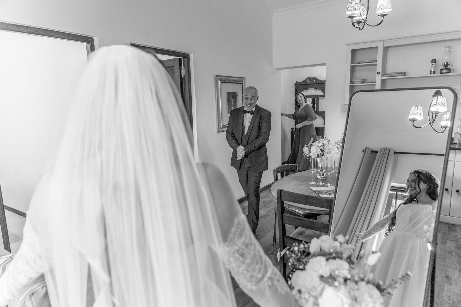 Black and white photo of a bride in veil holding bouquet as man in tuxedo watches her in a mirror.