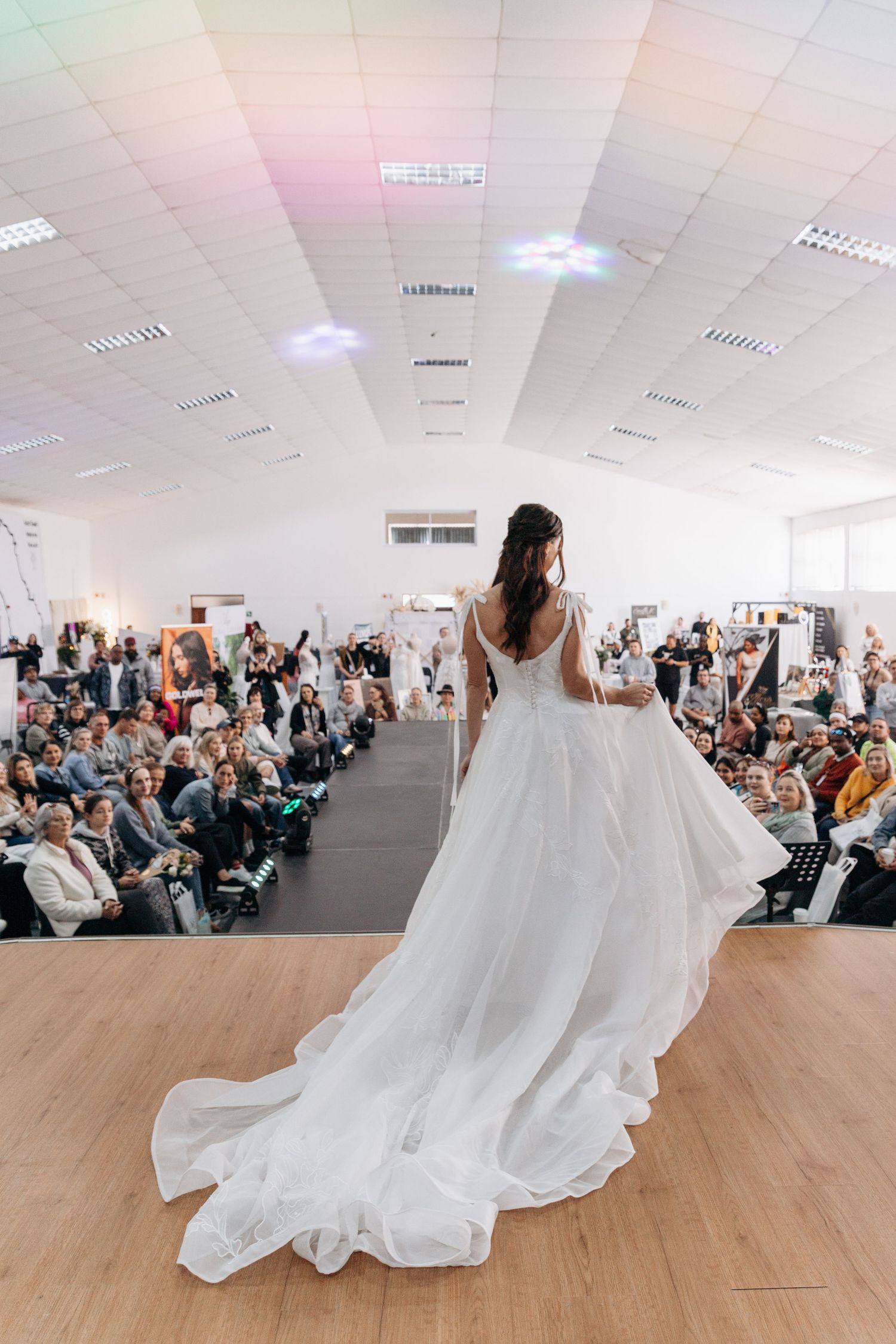 Bride in elegant white wedding gown with long train walks runway at bridal fashion show before seated audience.