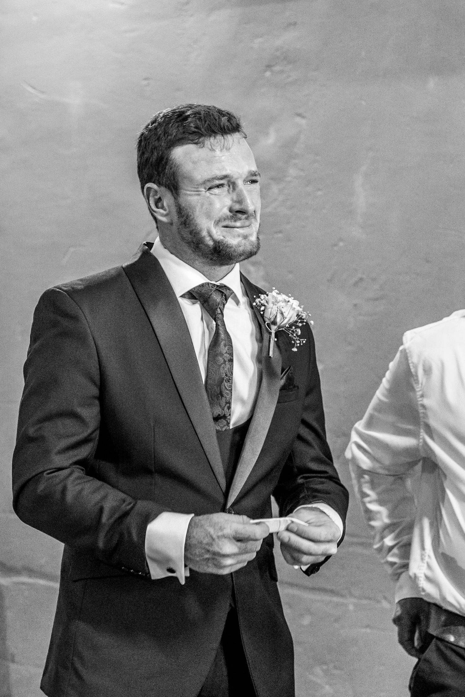 A groom in a black suit and floral boutonniere smiles during an outdoor wedding ceremony.