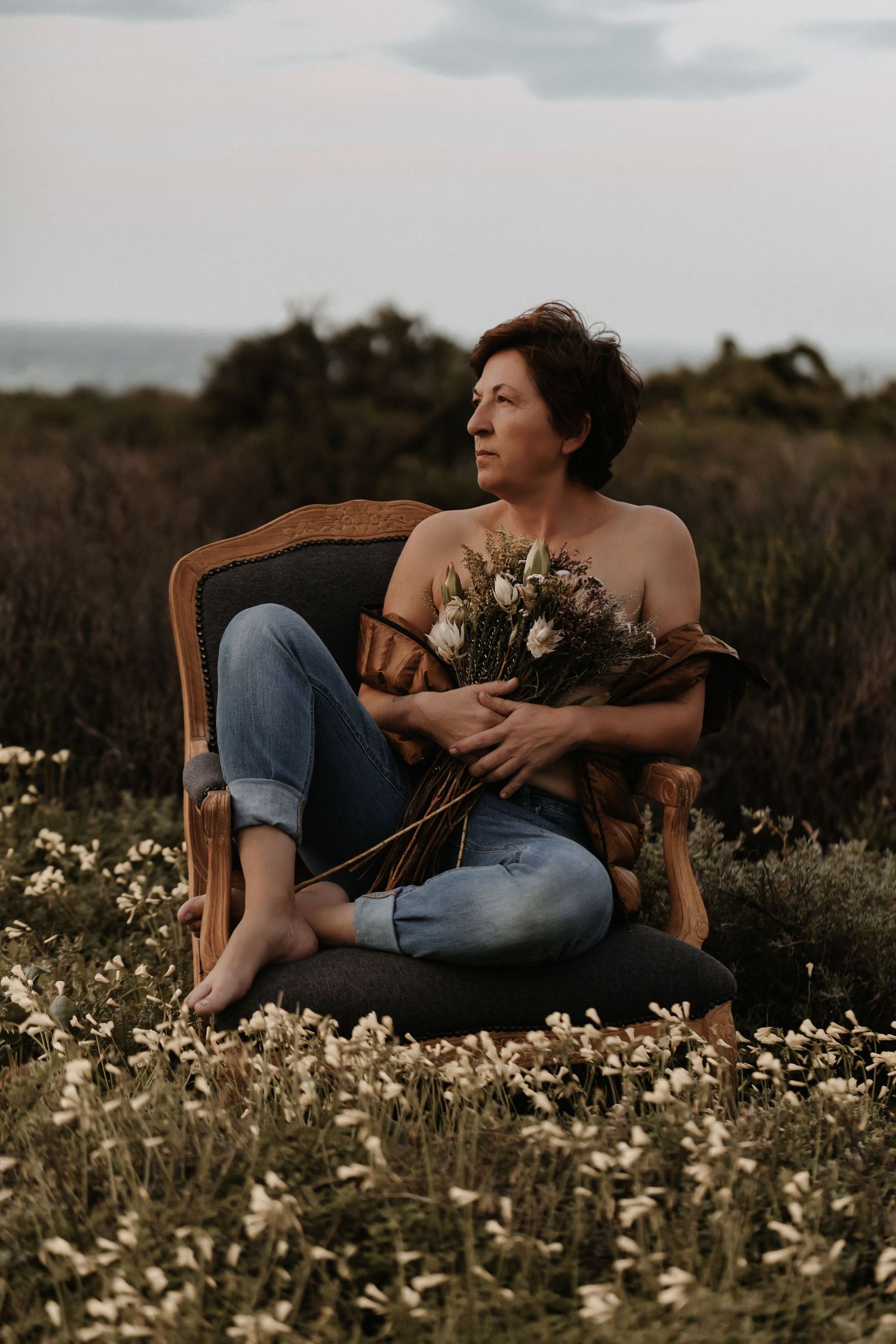 Person lounges in vintage wooden chair surrounded by delicate wildflowers in a dreamy summer meadow at dusk.