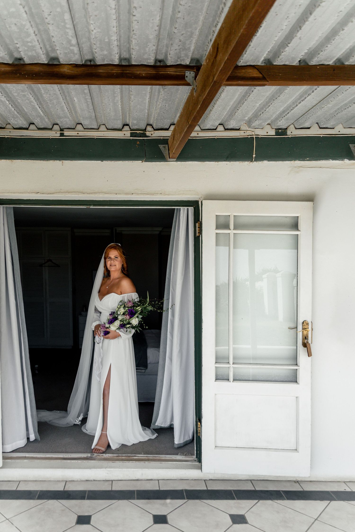 Bride in white off-shoulder gown holding bouquet standing in open doorway of rustic venue.
