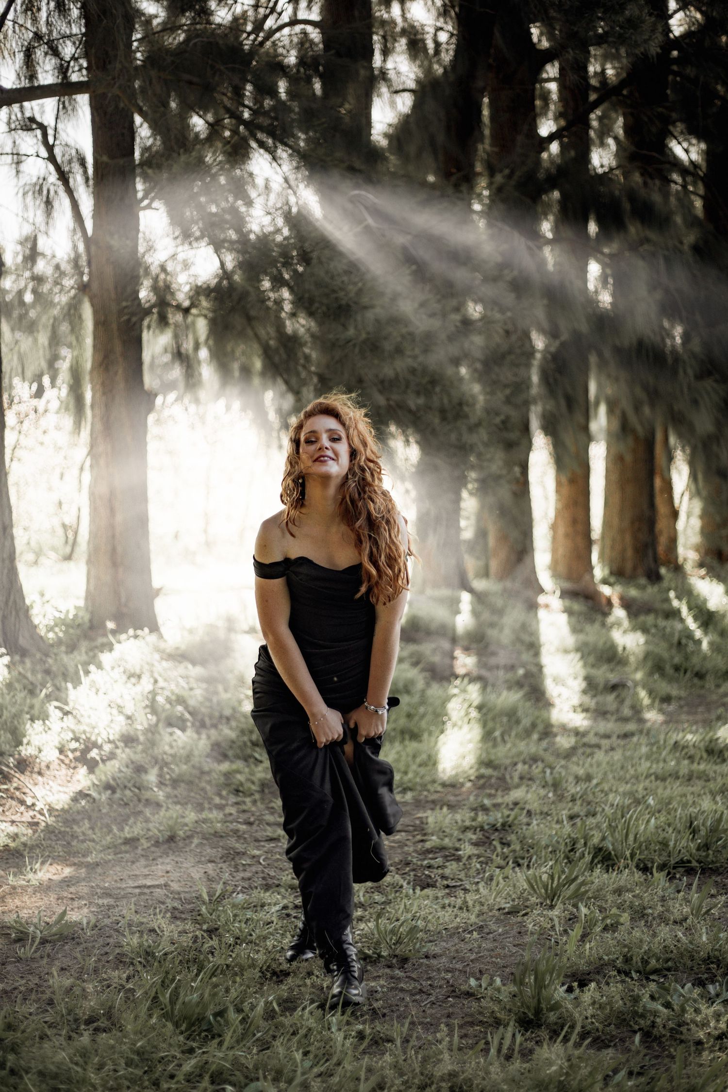 A person in a black off-shoulder dress stands in a misty forest with sunbeams streaming through the trees.