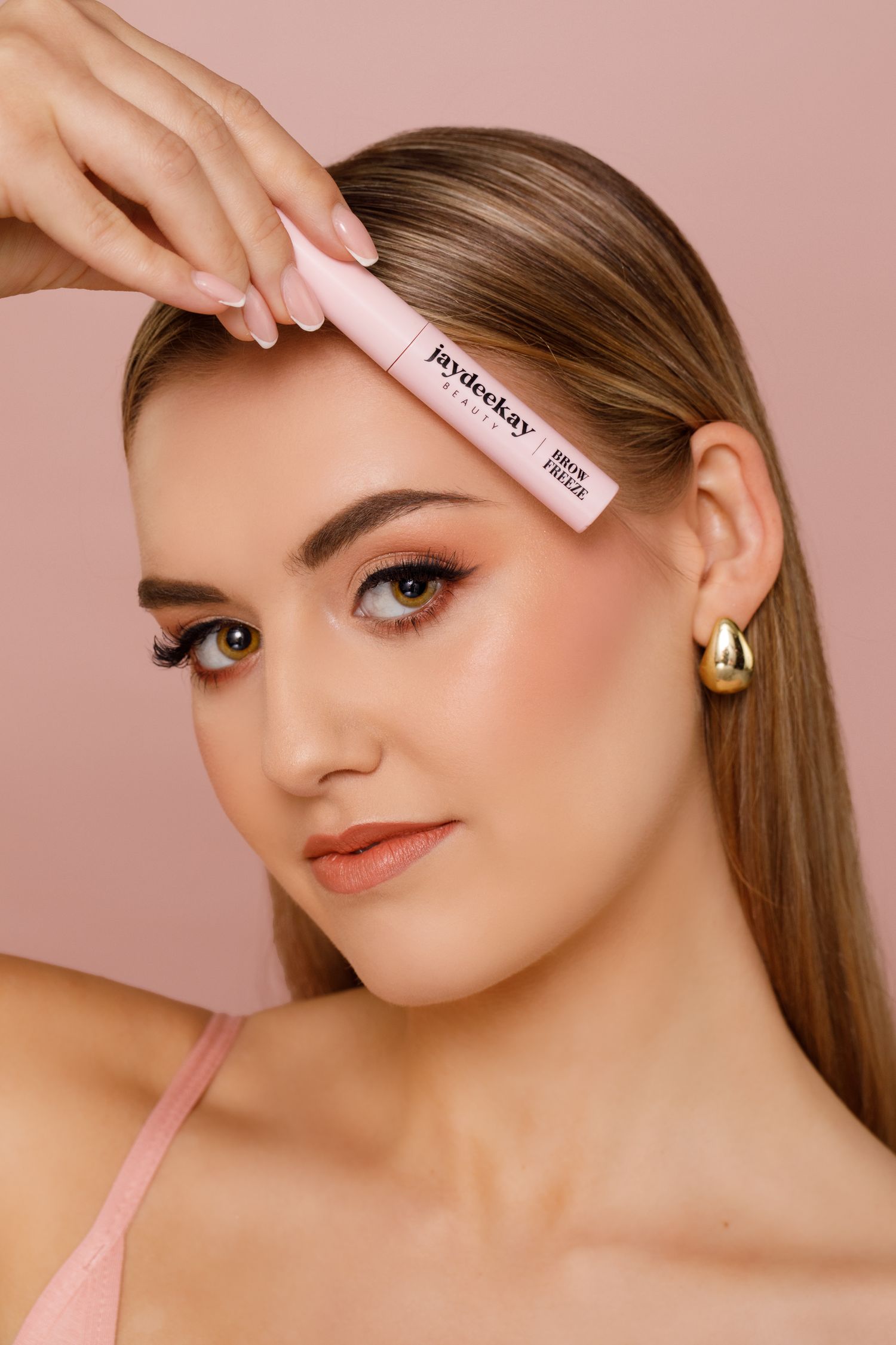 A beauty product demonstration shows application of concealer or highlighter on skin against a pink background.